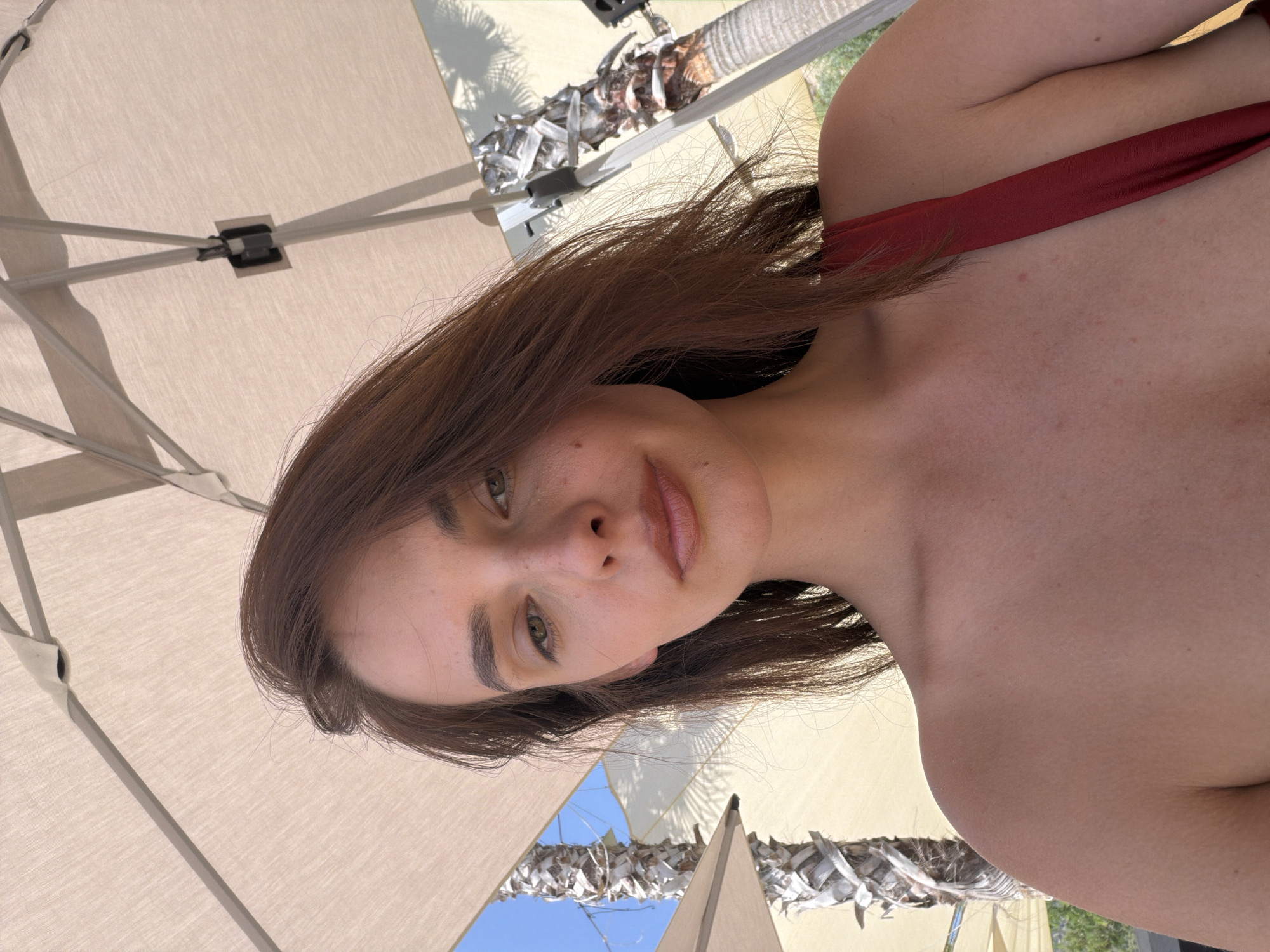 A woman is pictured from the chest up, in a selfie taken outdoors. She has long, brown hair and fair skin with freckles. She is wearing a red top. The background shows palm trees and what appears to be a beach umbrella, suggesting a warm, sunny climate. The overall scene is relaxed and casual, consistent with a vacation or leisure setting. No specific location in Fethiye, Turkey, is discernible from the image itself.