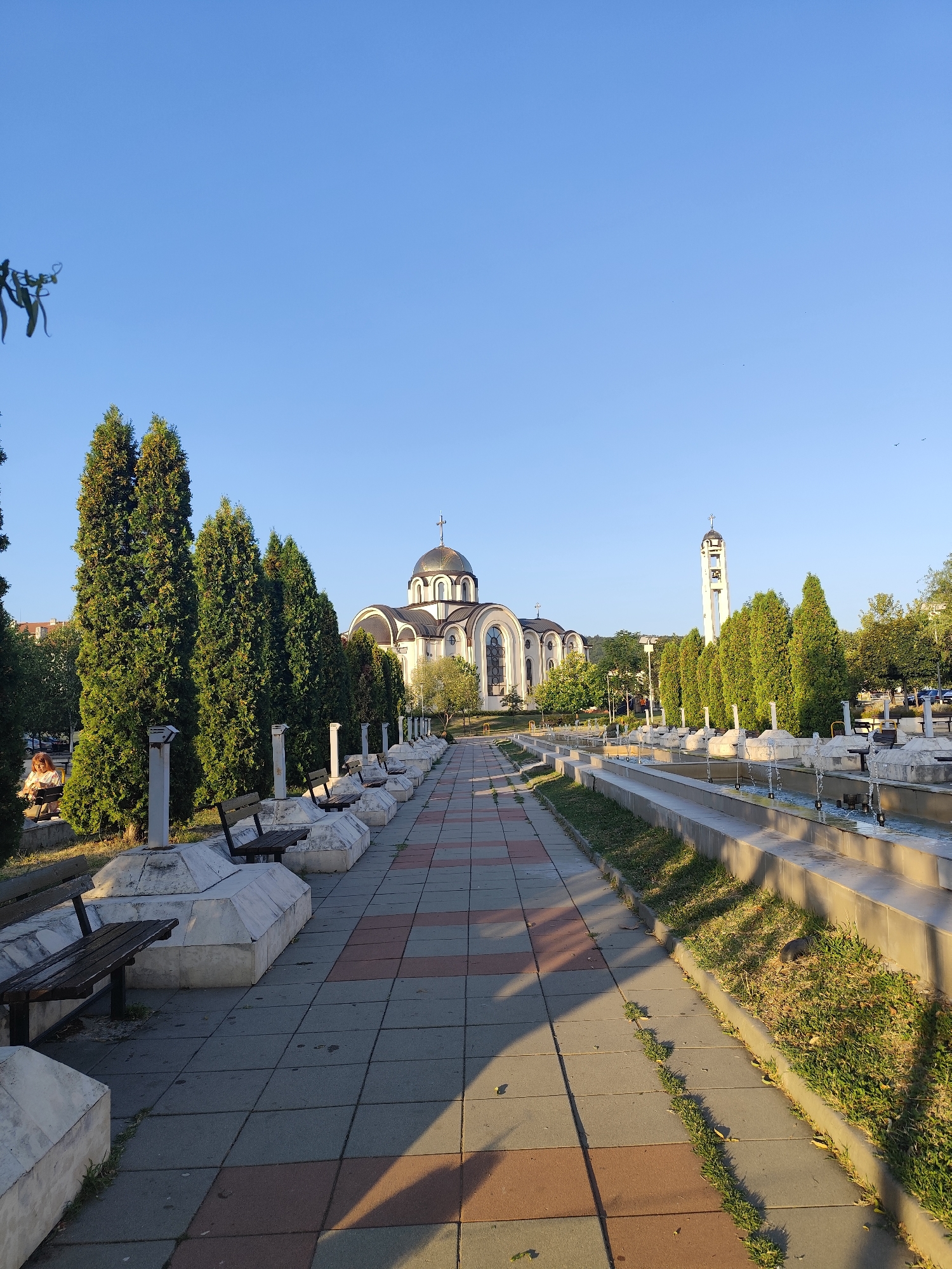 The image shows a wide paved walkway in an outdoor public space, likely a park or plaza, in the city of Montana, Bulgaria. The walkway is bordered by a row of evenly spaced, tall evergreen trees on the left and a tiered fountain system on the right. In the background, a large, white church with a golden dome and a tall bell tower are visible. Several benches are placed along the walkway, some occupied by individuals. The sky is clear and blue, indicating daytime.