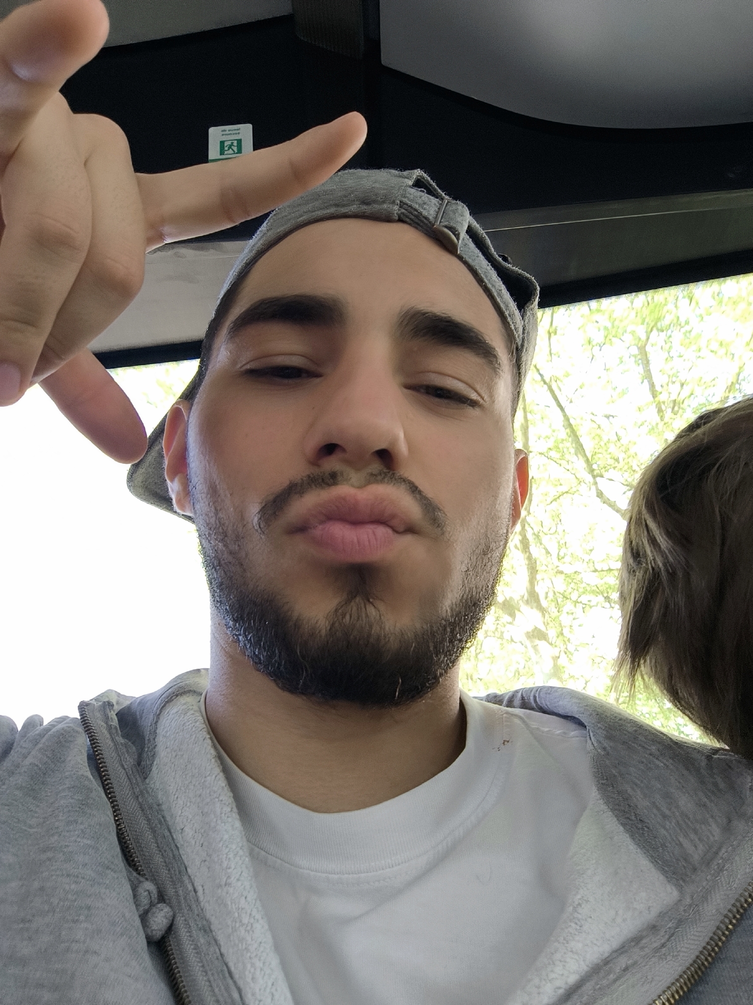 A man is captured in a close-up selfie, appearing to be inside a vehicle or building, with bright greenery visible through a window in the background. He has a short beard and mustache and is making a "kiss" face with pursed lips. He wears a grey baseball cap backwards and a grey zippered hooded sweatshirt over a white t-shirt. His left hand is raised to the upper left, making a gesture with his index and pinky fingers extended.
In the top-middle background, a small, green and white emergency exit sign is visible on a dark surface. The sign features a running man pictogram and the faintly discernible text "SORTIE DE SECOURS" (French for "Emergency Exit") and "EXIT". Bright natural light illuminates the scene, indicating daytime. Part of another person's brown hair is visible on the right side of the frame. The location context suggests this scene takes place in Strasbourg, France.