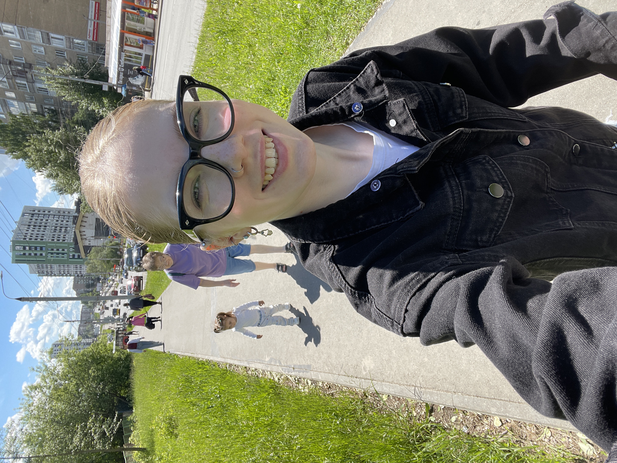 A selfie shows a fair-skinned person wearing large black glasses and a dark denim jacket. They are smiling with visible dental work. A nose ring is also apparent. In the background, pedestrians, including an adult in a purple shirt and a child in white, are walking on a sidewalk. Traffic is visible on the street alongside buildings, including a tall apartment complex. The scene is in daylight, with green grass bordering the sidewalk. The location is Izhevsk, Russia.