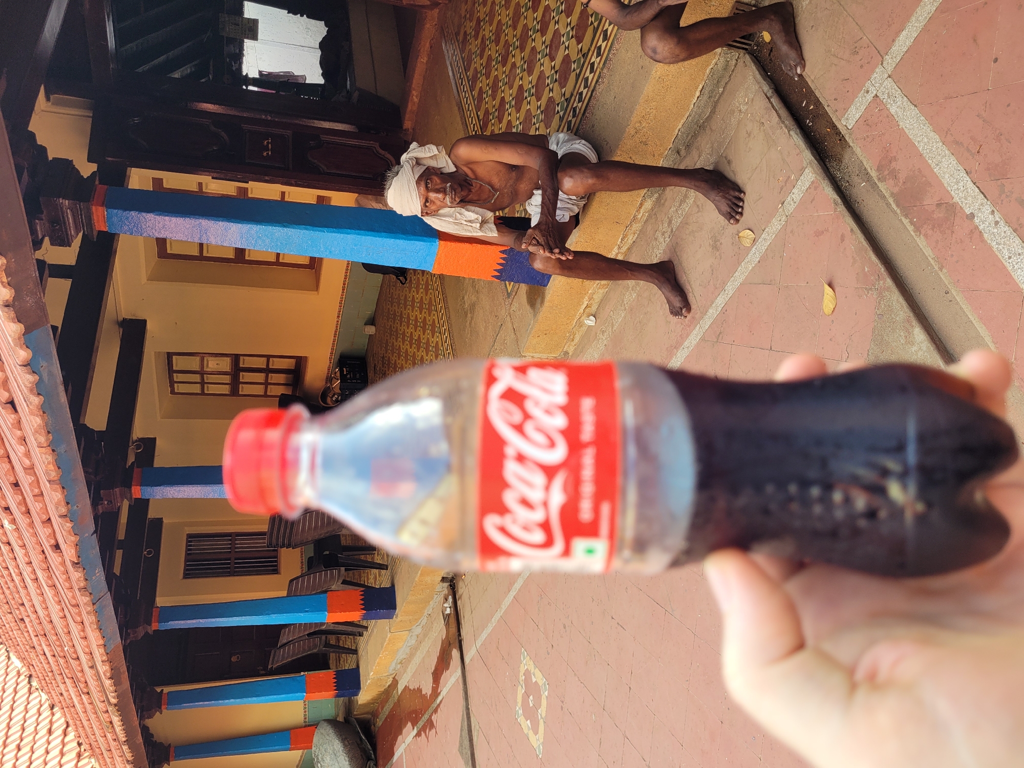 An eye-level shot captures a Coca-Cola bottle in the foreground, held in someone's hand, partially obstructing the view. In the background, an elderly man, possibly Indian, sits on the floor with his legs stretched out. He's wearing a white headwrap and appears to be resting. To the left, a building with blue and orange pillars is visible. The floor is patterned, suggesting it could be a courtyard or veranda. The scene suggests a location in India, possibly a rural or traditional setting.