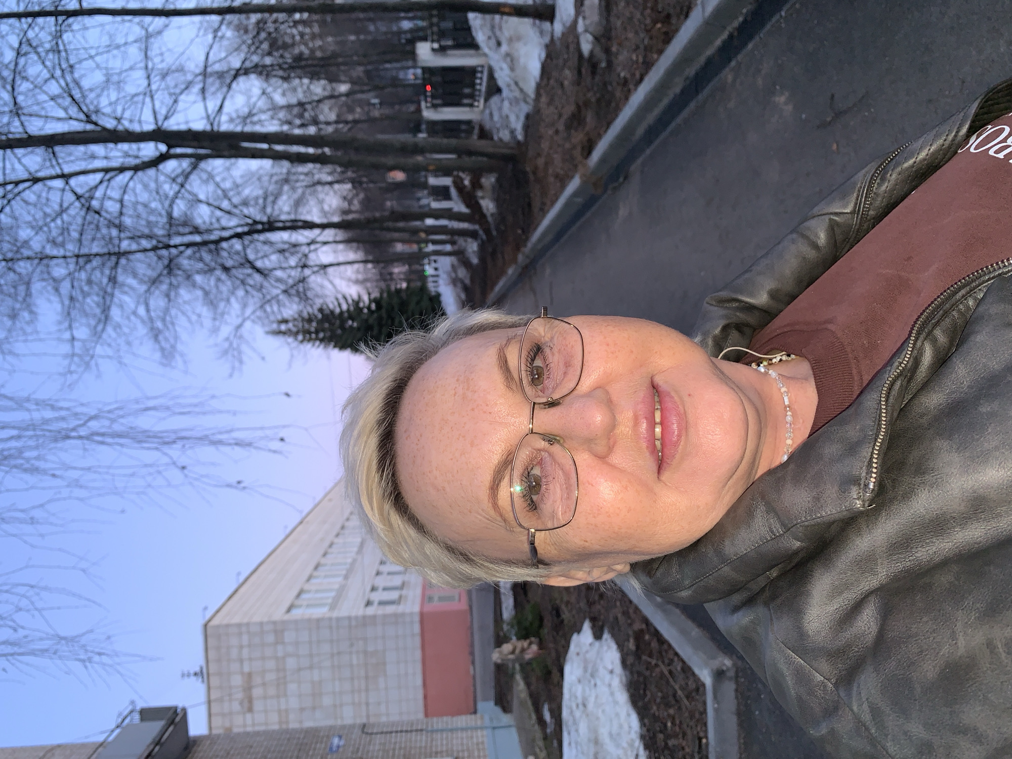 The image shows a middle-aged woman with short blonde hair and glasses, wearing a brown shirt and a dark leather jacket. She is smiling slightly at the camera. The background reveals a street scene in what appears to be the city of Chaikovsky, Russia, with leafless trees, a sidewalk, and a large building. Patches of snow are visible. The sky is pale blue, suggesting dusk or early morning. A fence and a street light can be seen further in the background.