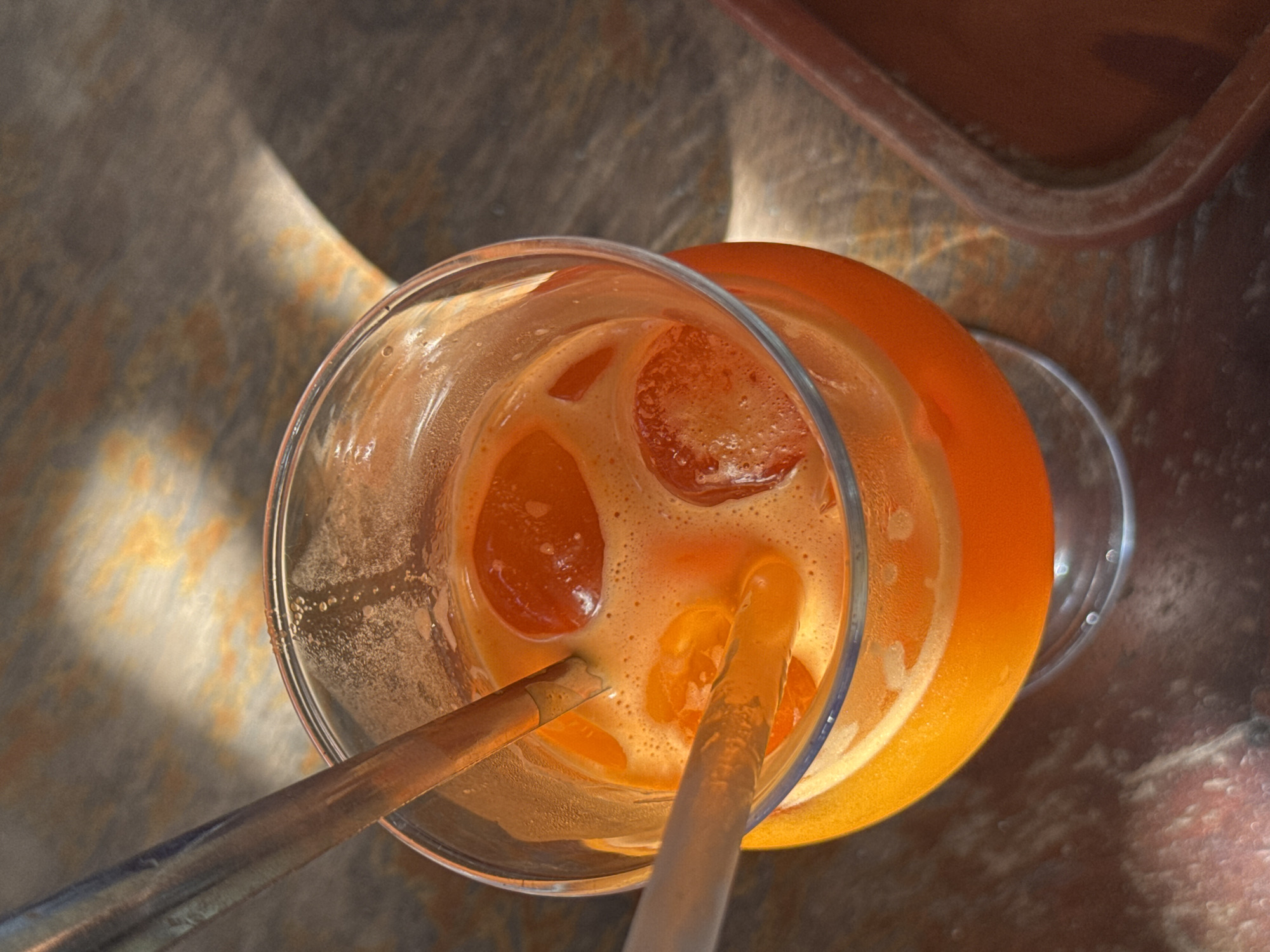 A close-up, overhead shot shows a clear glass filled with bright orange juice and ice cubes. Two metal straws are inserted into the juice. The glass sits on a weathered, wooden surface, possibly a table. The lighting creates dappled shadows across the scene, suggesting it is outdoors or near a window. A portion of a reddish-brown, textured object, potentially a planter or bowl, is visible on the right side of the frame. The image captures a refreshing beverage, likely enjoyed in a relaxed setting within Nam Nha Trang Ward, Vietnam.
