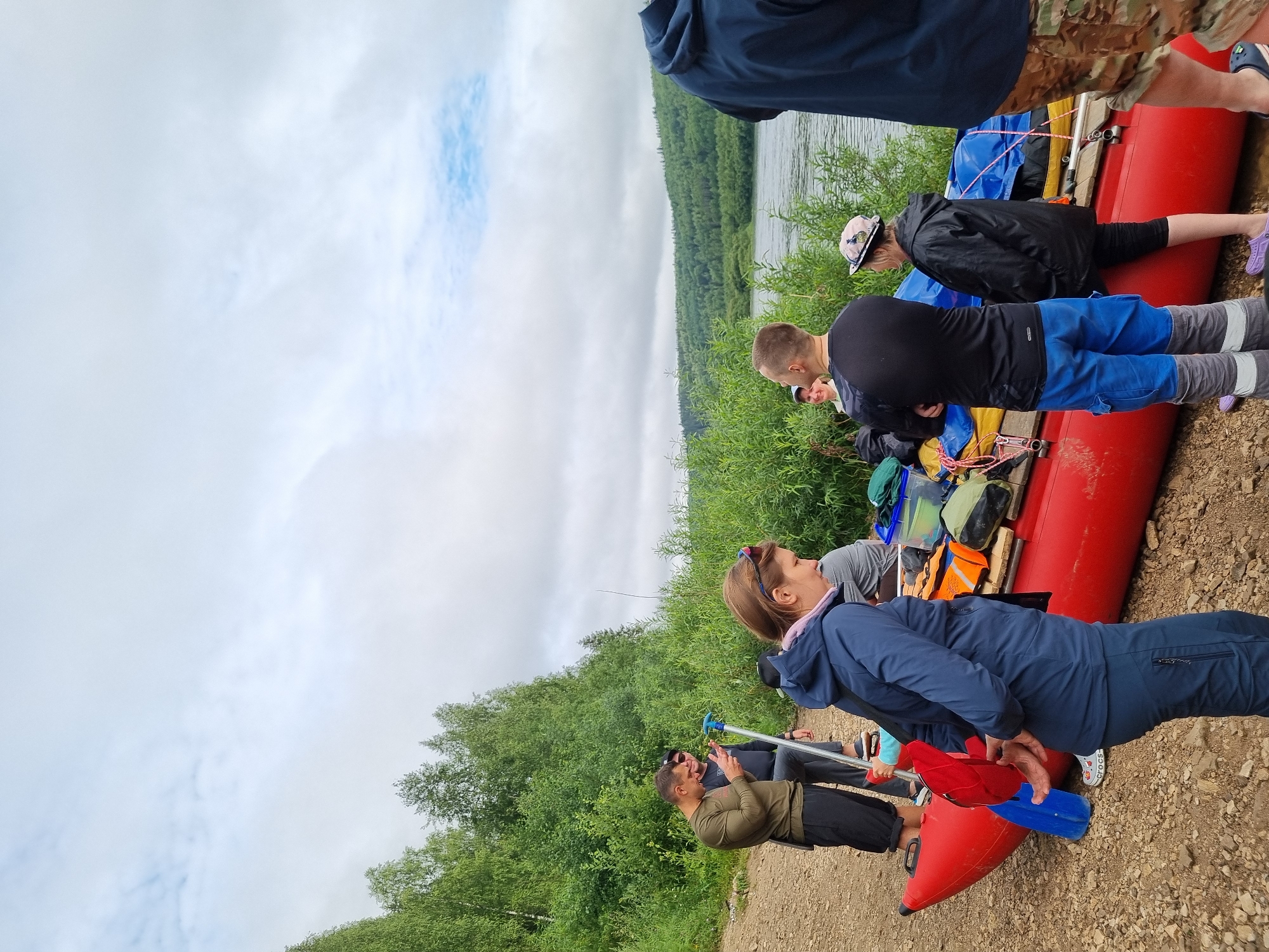A group of people are gathered on a rocky shoreline next to a bright red raft. The raft is loaded with various gear, including bags and life vests. Dense green foliage and trees form a backdrop, with a wide river flowing behind them. The sky is overcast with scattered clouds. The people appear to be preparing for or finishing an outdoor activity, likely related to water sports given the presence of the raft. The scene is set in a natural, possibly wilderness area near Visherogorsk, Russia.