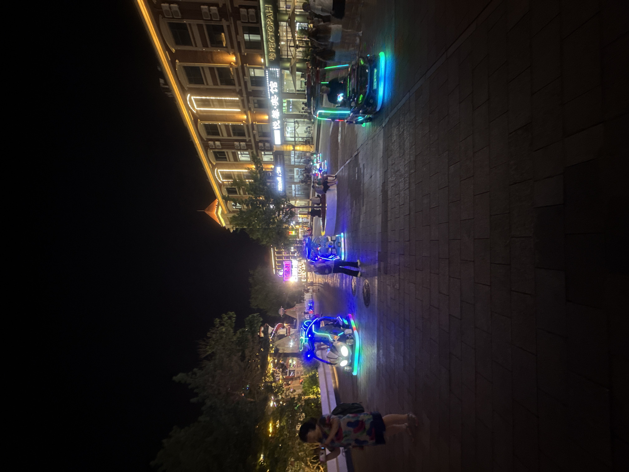 This is a nighttime, eye-level, medium shot of a pedestrian street in Qinhuangdao, China, likely in the Nandaihe tourist area, known for its nightlife and entertainment. A woman in a colorful dress is standing on the left side of the frame, holding a smartphone. In the foreground, several illuminated, miniature, futuristic-looking vehicles with colorful LED lights are parked on a brick-paved walkway. These vehicles appear to be small, electric-powered rides designed for entertainment. In the background, there are trees with decorative lights, buildings with illuminated windows and signage, and other pedestrians. One building in the background has signage in Russian, "РЕСТОРАН" (Restoran), indicating a possible Russian cultural influence or presence in the area. The overall atmosphere is festive and lively, typical of a commercial or entertainment district at night.