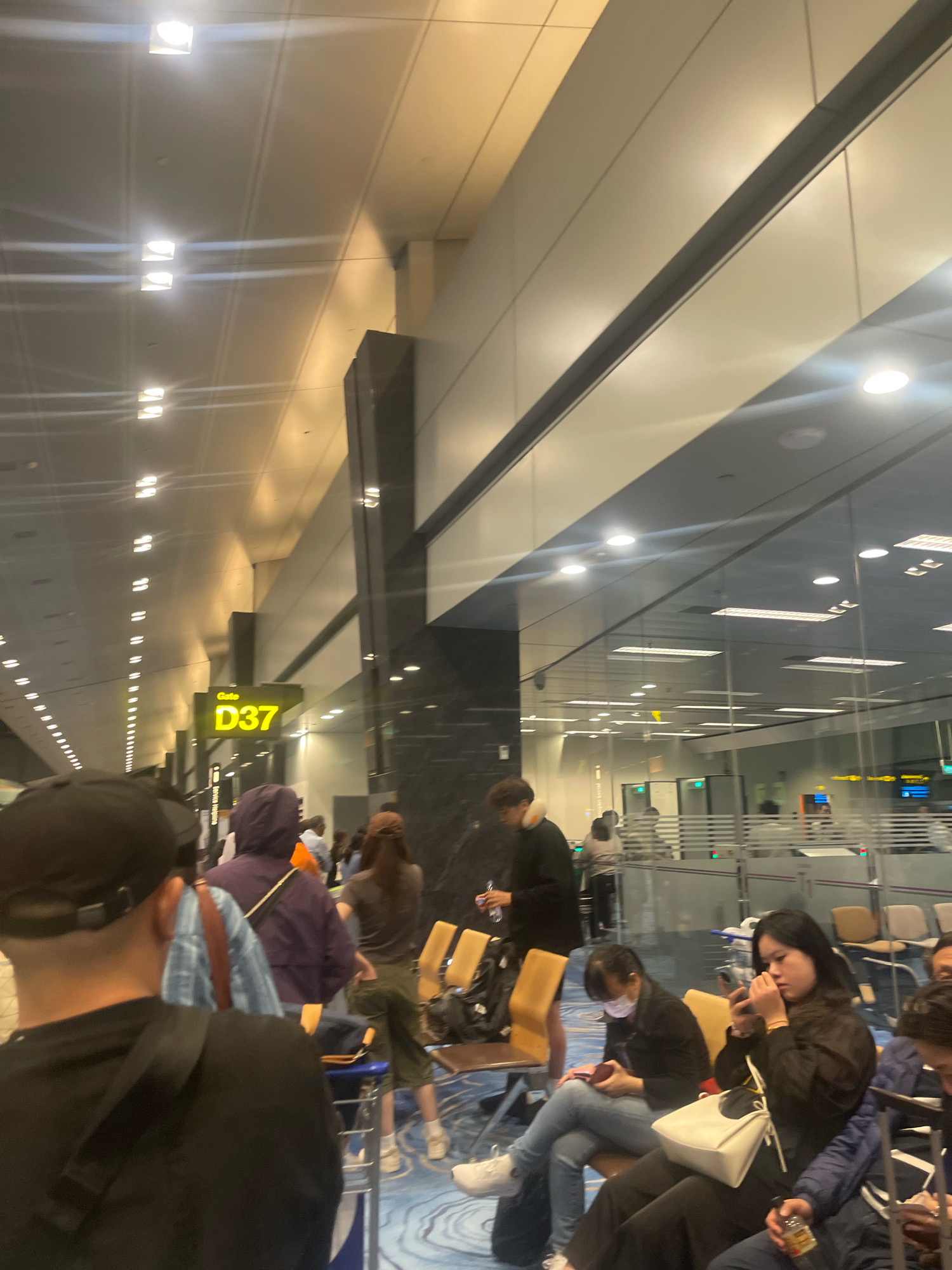 The image depicts a scene inside an airport terminal, likely in Singapore, Singapore, as indicated by a visible "Gate D37" sign. Several individuals are waiting in a queue or seated on chairs. The environment appears to be a modern airport with bright lighting, reflective surfaces, and glass partitions.
In the foreground, a person wearing a black t-shirt and a black baseball cap is visible from the back, facing away from the camera. To their left, another person in a purple hoodie is seen from the back, also waiting. Further back, a woman in a dark jacket is seated, looking at her phone. Other individuals are seated and standing, some engaged with their phones or bags.
The airport seating area is visible, with chairs arranged in rows. Luggage and bags are scattered around, consistent with an airport setting. The overall atmosphere suggests a typical waiting area before boarding a flight. The lighting is a mix of overhead fluorescent lights and some softer ambient light, creating a well-lit but somewhat sterile environment. The floor has a patterned design, and the walls appear to be a combination of light-colored panels and dark, possibly stone or metallic, sections.