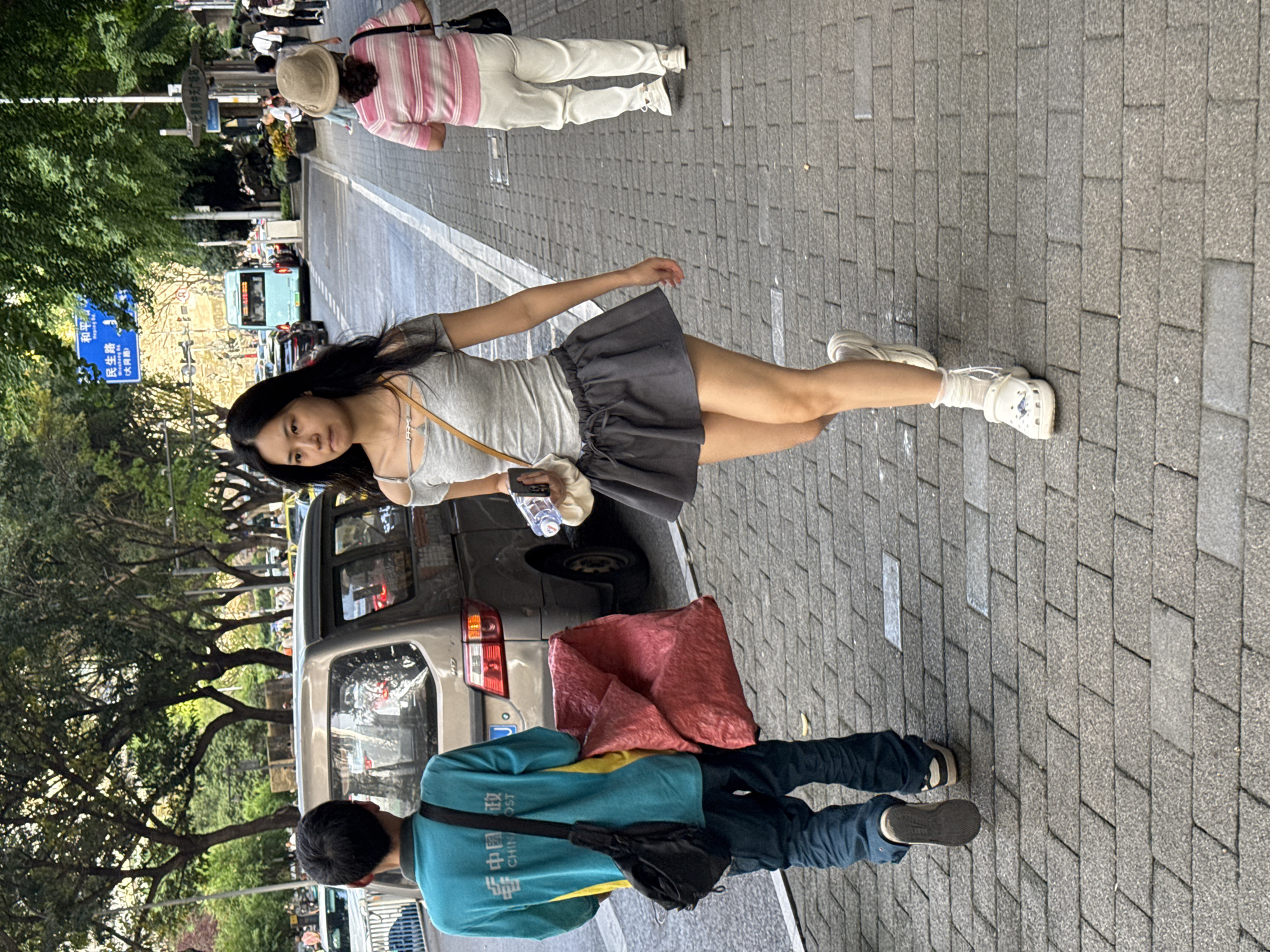 A street scene in Yuzhong District, China, shows a young woman with long black hair wearing a gray top and dark gray skirt, light-colored socks and white Crocs, standing on a paved street. She holds a water bottle and a small beige bag. Behind her, a gray car is parked along the curb. Next to the car, a delivery person wearing a blue jacket and dark pants stands with a red package next to them. In the background, there are trees, a street sign with Chinese characters, and pedestrians.