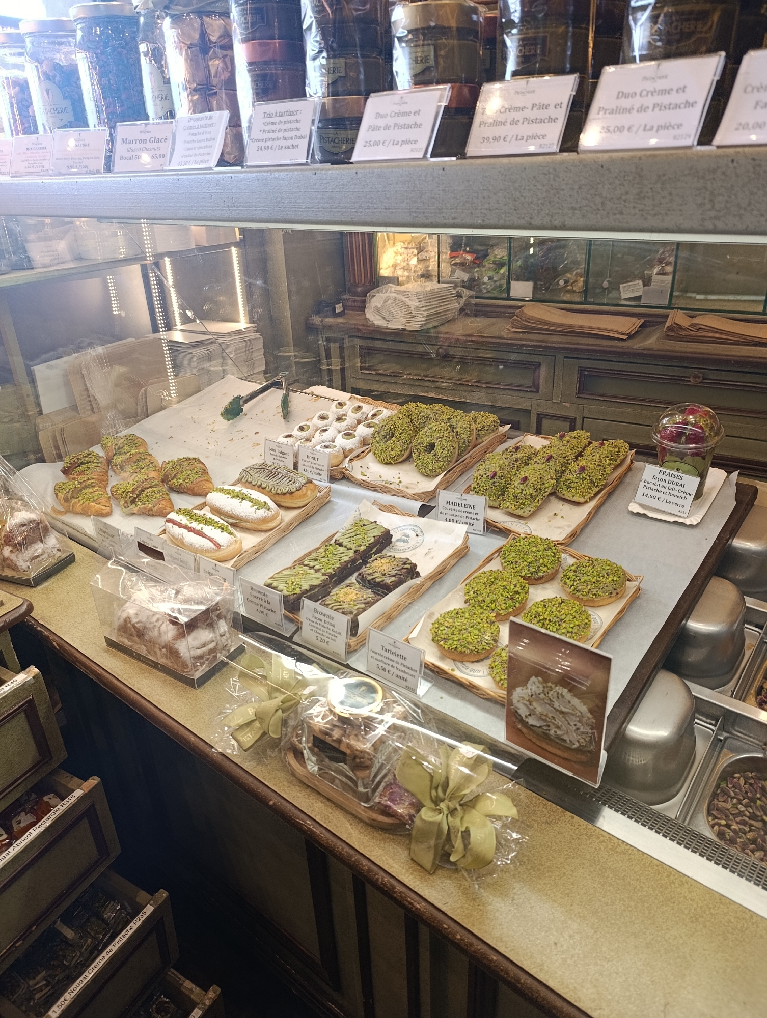 The image shows a display case in a pastry shop, likely located in Paris, France. The case is filled with various pastries and sweets, including croissants, donuts, brownies, madeleines, and tarts, many of which are decorated with pistachios. Price tags are displayed for each item. Jars of spreads and preserved fruits are visible on the shelves above the display case. Below the display case are drawers containing additional sweets, such as nougat and fruit preserves. The pastries and sweets are beautifully arranged and appear to be fresh.