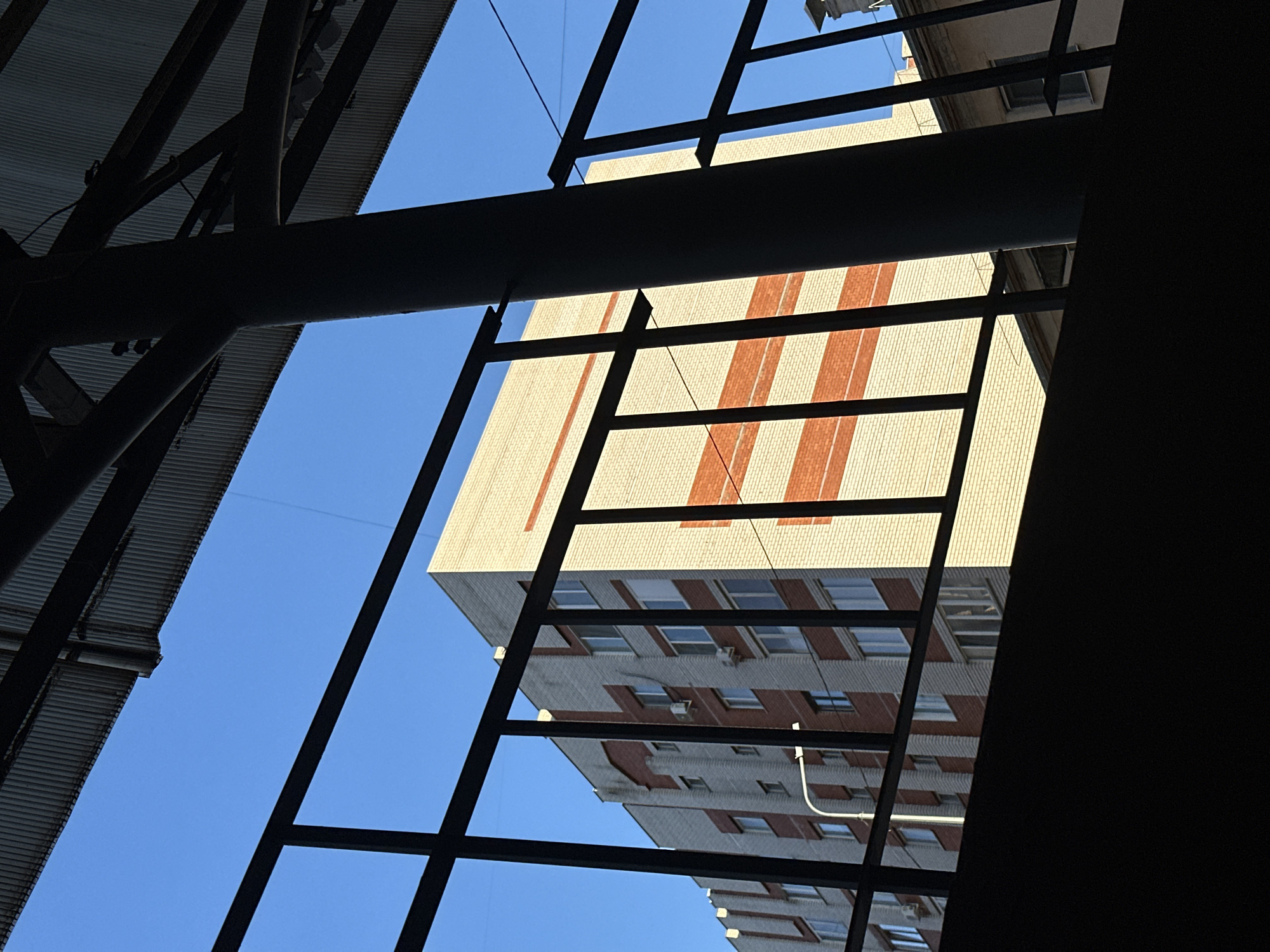 A low-angle shot looking up at a building framed by metal construction. The sky is blue. The building has a brick facade with alternating bands of light and reddish-brown brick. Windows are visible on the lower portion of the building. The metal construction is black and forms a grid-like pattern. The setting is Kazan, Russia.