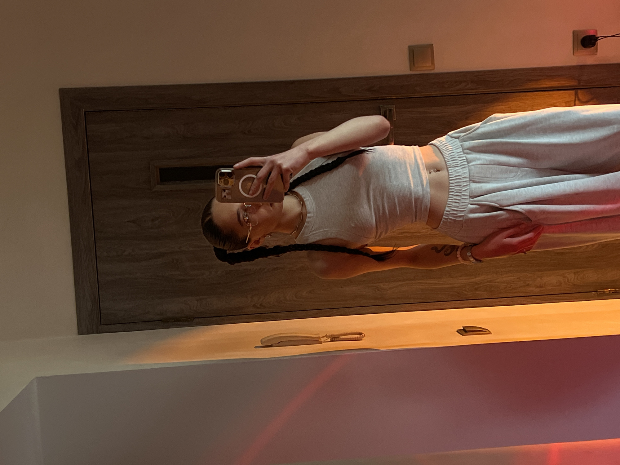 A person is taking a mirror selfie in what appears to be a bedroom or hallway. The person has dark hair styled in two braids and is wearing glasses. They are dressed in a light-colored tank top and sweatpants. A smartphone is held up to take the photo, reflecting in the mirror. Behind the person, a wooden door is visible. In the foreground, on a shelf-like surface, is a landline telephone. The lighting suggests it is likely evening or indoors with artificial light. There are electrical outlets visible on the wall. The image also shows colorful light reflections on the surfaces.