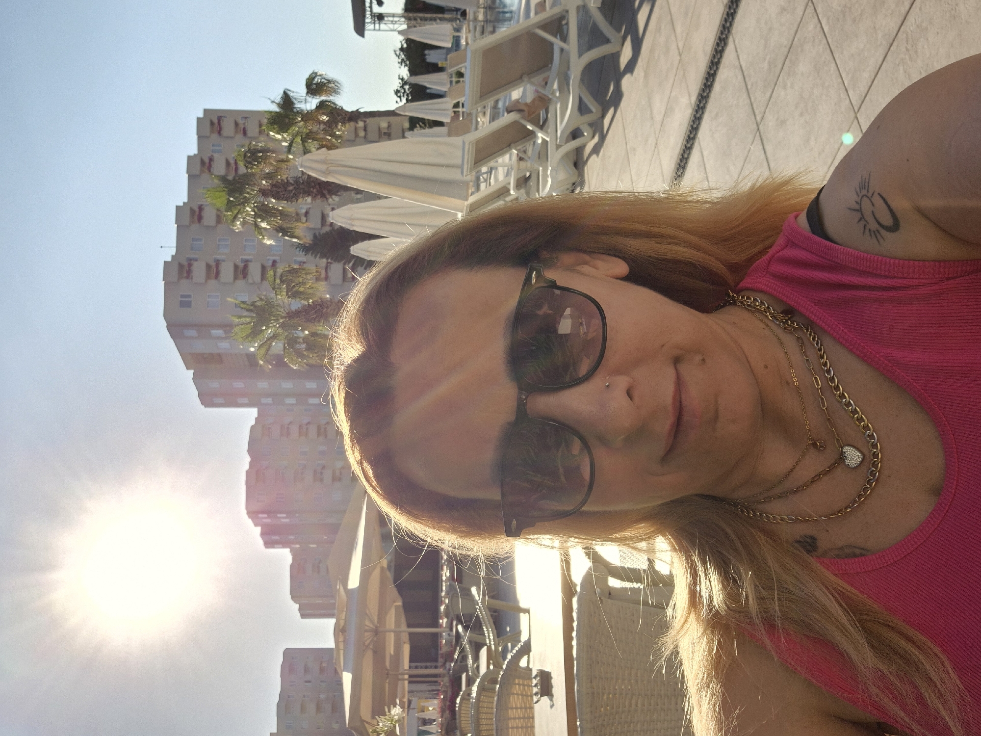A woman with blonde hair and sunglasses is taking a selfie outdoors. She is wearing a pink tank top and multiple necklaces. There is a tattoo on her left shoulder. In the background, there are buildings, palm trees, lounge chairs, and umbrellas, suggesting a resort or hotel setting. The sun is shining brightly in the sky, creating lens flare in the image.