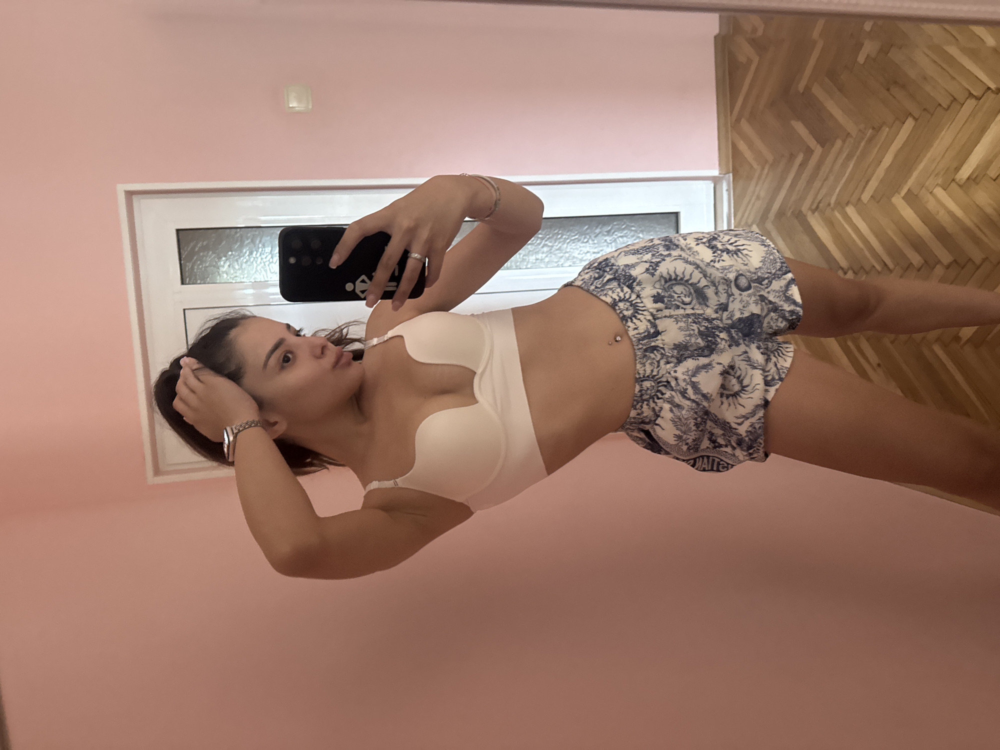 A young woman is taking a mirror selfie indoors, captured from a horizontally rotated perspective with her head on the left. She is wearing a white bra and blue and white patterned shorts, featuring sun-like and abstract designs. A belly button piercing is visible. She wears a ring on her right ring finger and a bracelet on her left wrist. Her right hand holds a dark smartphone, with a light-colored emblem on its back case, positioned to photograph her reflection. Her left hand is near her temple, adjusting her dark hair, and she has a playful expression with her tongue slightly out.
The background, seen through the mirror's reflection, reveals a room with light pink walls and a wooden floor laid in a herringbone pattern. A white-framed window with frosted or obscured glass is visible behind her. A small, rectangular light switch or fixture is also present on the pink wall. The setting is an indoor space, likely a private room. There is no discernible text visible on her clothing or phone. The location context for the image is Plovdiv, Bulgaria.