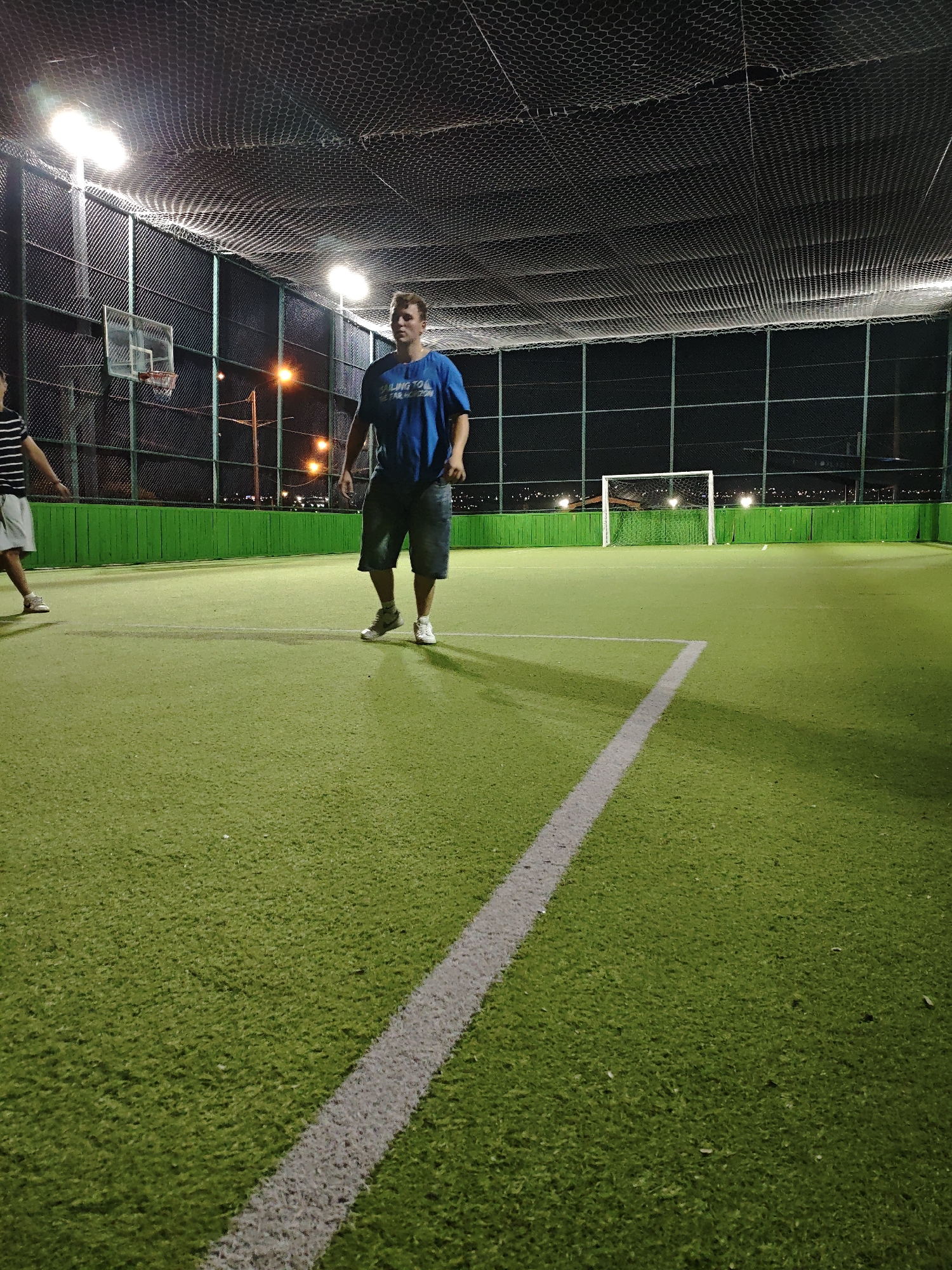 An exterior shot captures two males on a brightly lit, artificial turf soccer field at night. The field has white markings and a green perimeter fence. A white soccer goal is visible in the background, as is a basketball hoop. The man in the foreground, wearing a blue t-shirt and dark shorts, stands looking forward. To his left, another male in a striped shirt and light shorts is walking away from the camera. The scene is enclosed by a netted ceiling, suggesting an indoor or covered outdoor facility. Lights illuminate the field from above.