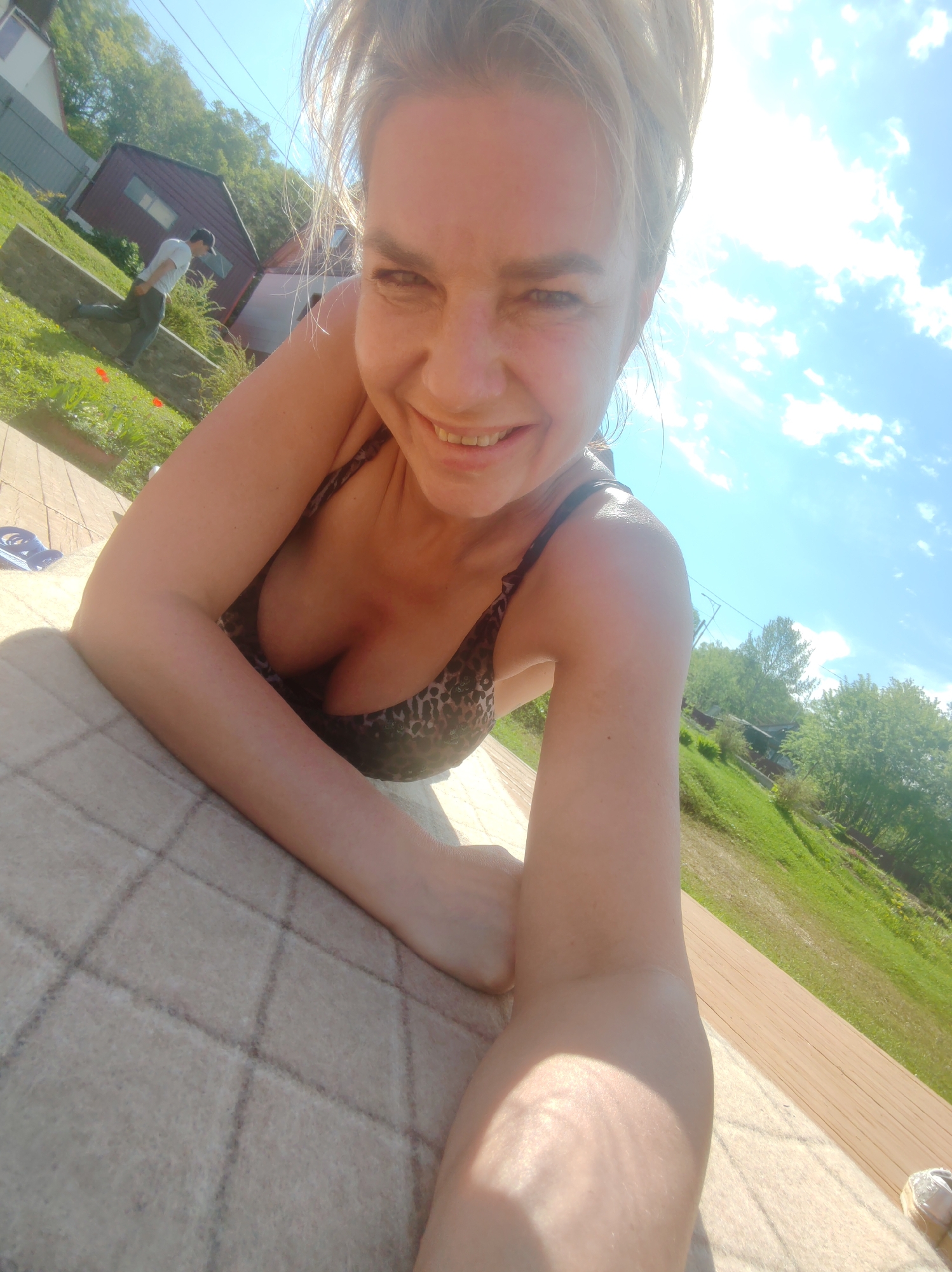 A blonde woman is lying on a textured surface outdoors, looking into the camera and smiling. She is wearing a leopard-print bikini top. In the background, a man in a white t-shirt and dark pants is walking away from the camera. The environment appears to be a grassy hillside with trees and some buildings. The sky is blue with scattered clouds. The location is Paratunka, Russia.