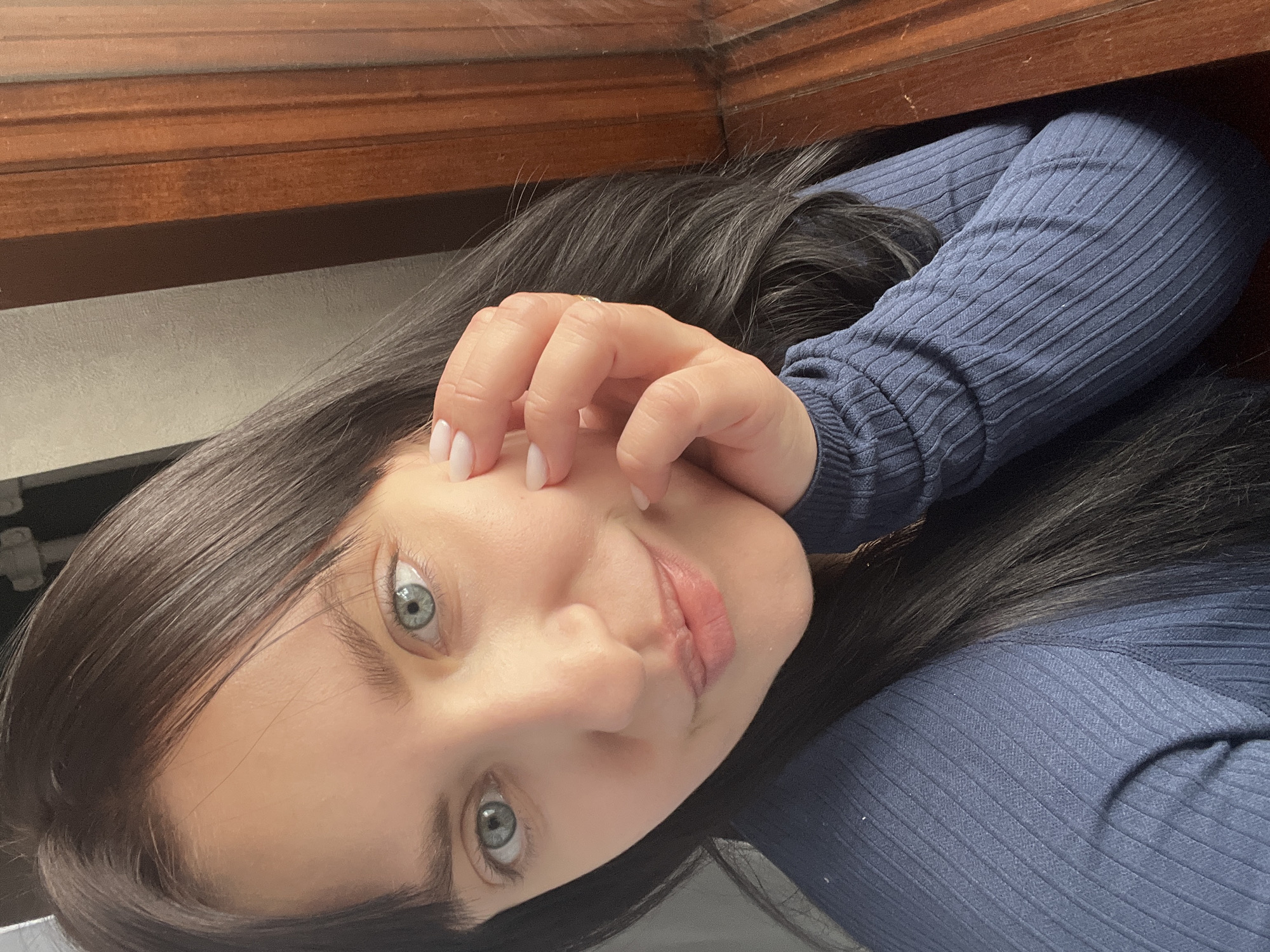 The image features a young woman with dark hair and striking light blue-grey eyes, looking directly at the camera with a calm expression. She appears to be lying down or reclining, with her head tilted, suggesting a selfie taken from an overhead perspective. Her right hand, adorned with light-colored nail polish and a ring on the ring finger, is gently positioned near her cheek and temple. She is wearing a close-fitting, long-sleeved top made of dark blue ribbed fabric, which is visible across her chest and arm.
The background suggests an indoor setting. To the left of her head (as oriented in the image) is a prominent dark brown wooden surface, possibly a shelf or part of a furniture piece. Below this wooden element, a lighter, plain wall or surface is visible. The lighting is soft and even, highlighting her facial features. No visible text is present in the image. The scene is set in Марковский, Russia.