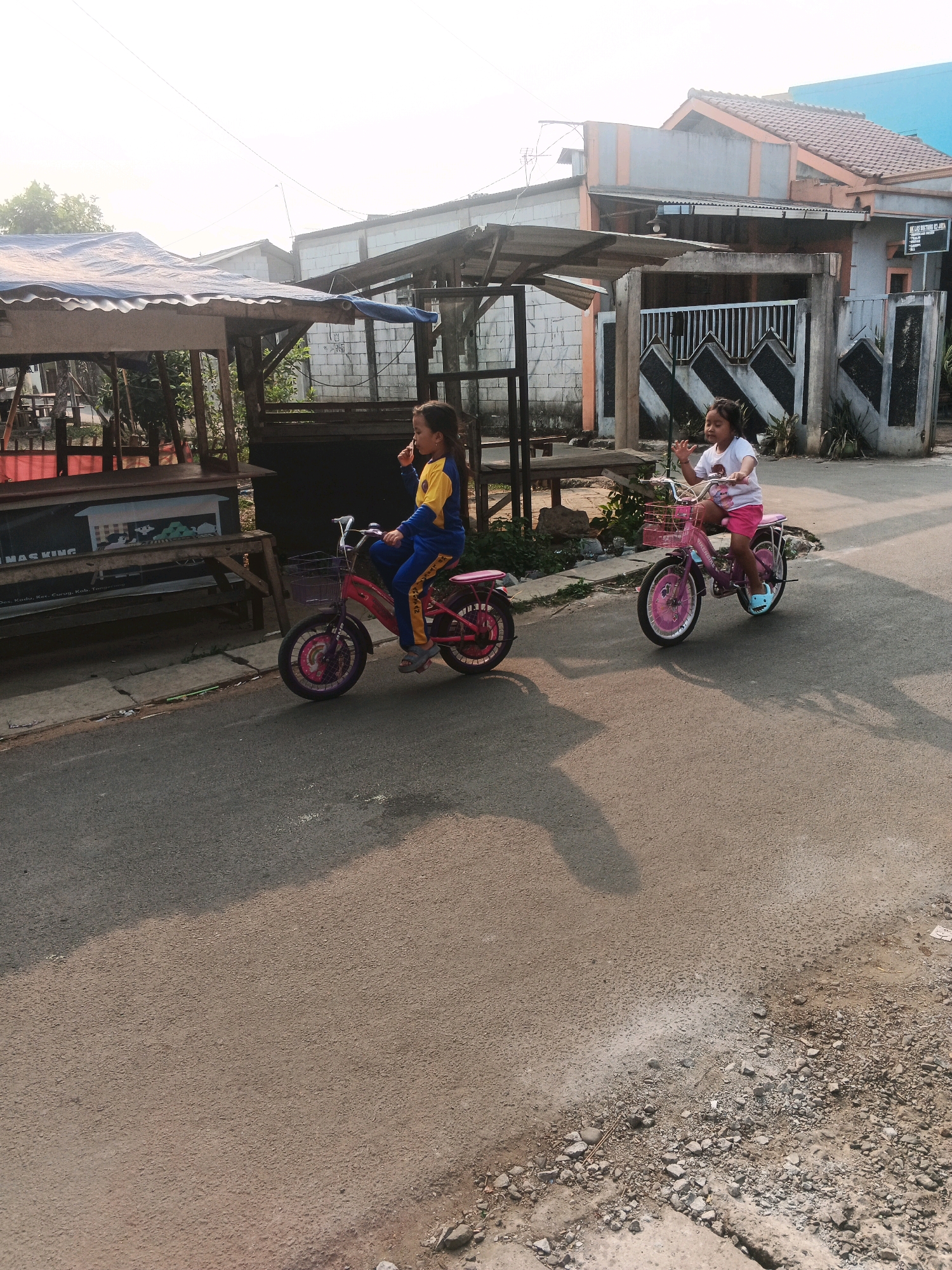 Two young girls ride pink bicycles on a paved street in front of houses in Kadu, Indonesia. The girl on the left, wearing a yellow and blue tracksuit, rides a bicycle with a purple basket. She is looking up and to the right. The girl on the right, wearing a pink shirt and shorts, rides a bicycle with a pink basket. She is looking forward and has her right hand raised. Both bicycles have training wheels. The street is lined with houses and a small market stall. The sky is bright and overcast.