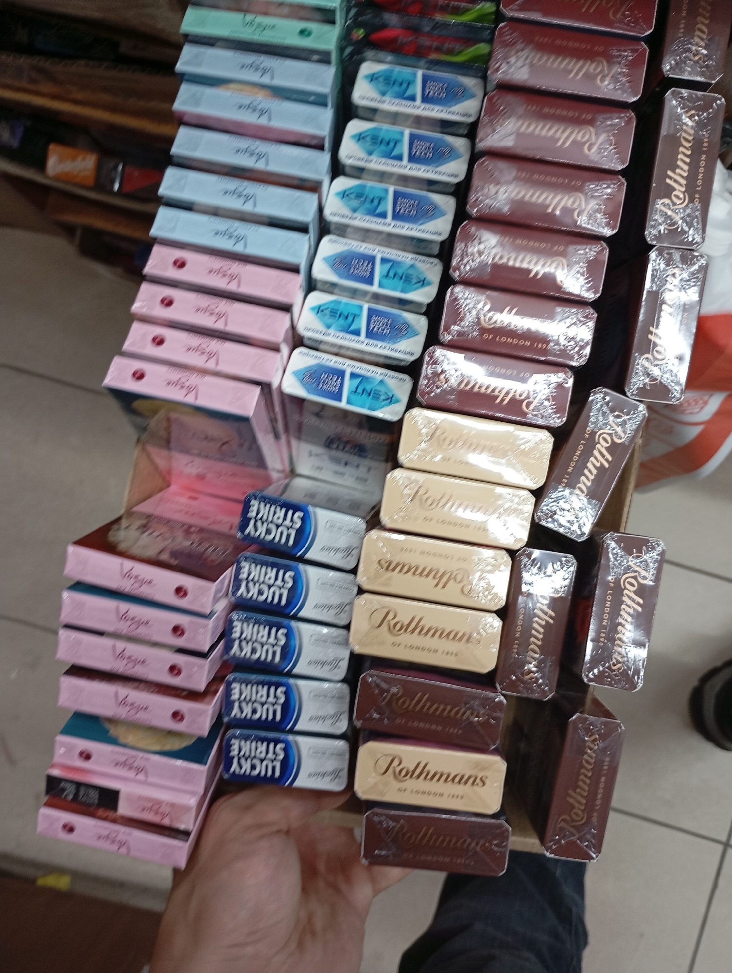 The image shows a retail display of tobacco products, primarily cigarettes, in a store. The products are stacked in neat rows and columns, with various brands and packaging designs visible. Some of the visible brands include "Vogue," "Kent," "Lucky Strike," and "Rothmans." The products appear to be shrink-wrapped. The background suggests an indoor environment, possibly a convenience store or a tobacco shop, with shelves and other merchandise faintly visible. The lighting is moderate, and the overall impression is of an organized inventory display. The image does not provide enough information to determine its specific location in Moscow, Russia, or any details about people, actions, or interactions.