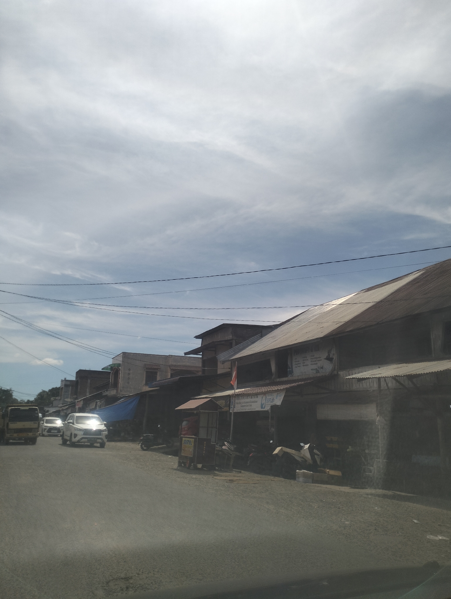 This image captures a street scene in Kruengmanyang, Indonesia, on a cloudy day. The sky is filled with wispy, light-colored clouds against a pale blue background, suggesting either early morning or late afternoon light. The foreground shows a paved road with a slight texture, indicating it might be a bit worn.
Along the right side of the road, a row of buildings with corrugated metal roofs lines the street. These buildings appear to be small shops or businesses, some with open fronts and awnings. One building has a sign with text that is partially obscured but includes what looks like "PHOTOCOPY & SPA" in English, and further text below in what appears to be Indonesian. Another sign reads "Sufiah". There are several parked motorcycles and what looks like some stacked materials outside one of the shops.
On the left side of the road, several vehicles are visible. A yellow truck is in the distance, followed by two white cars. The car closest to the viewer is a modern SUV, with its headlights on. The presence of these vehicles and the road suggest active local transportation.
The overall atmosphere is that of a typical day in a small town or village, with commerce and daily life unfolding. The weather seems mild, with the clouds diffusing the sunlight. There are no people prominently featured in the image, but the activity of vehicles and businesses implies human presence and ongoing routines. The setting is distinctly Indonesian, with the architectural style of the buildings and the presence of a truck commonly seen in Southeast Asia.