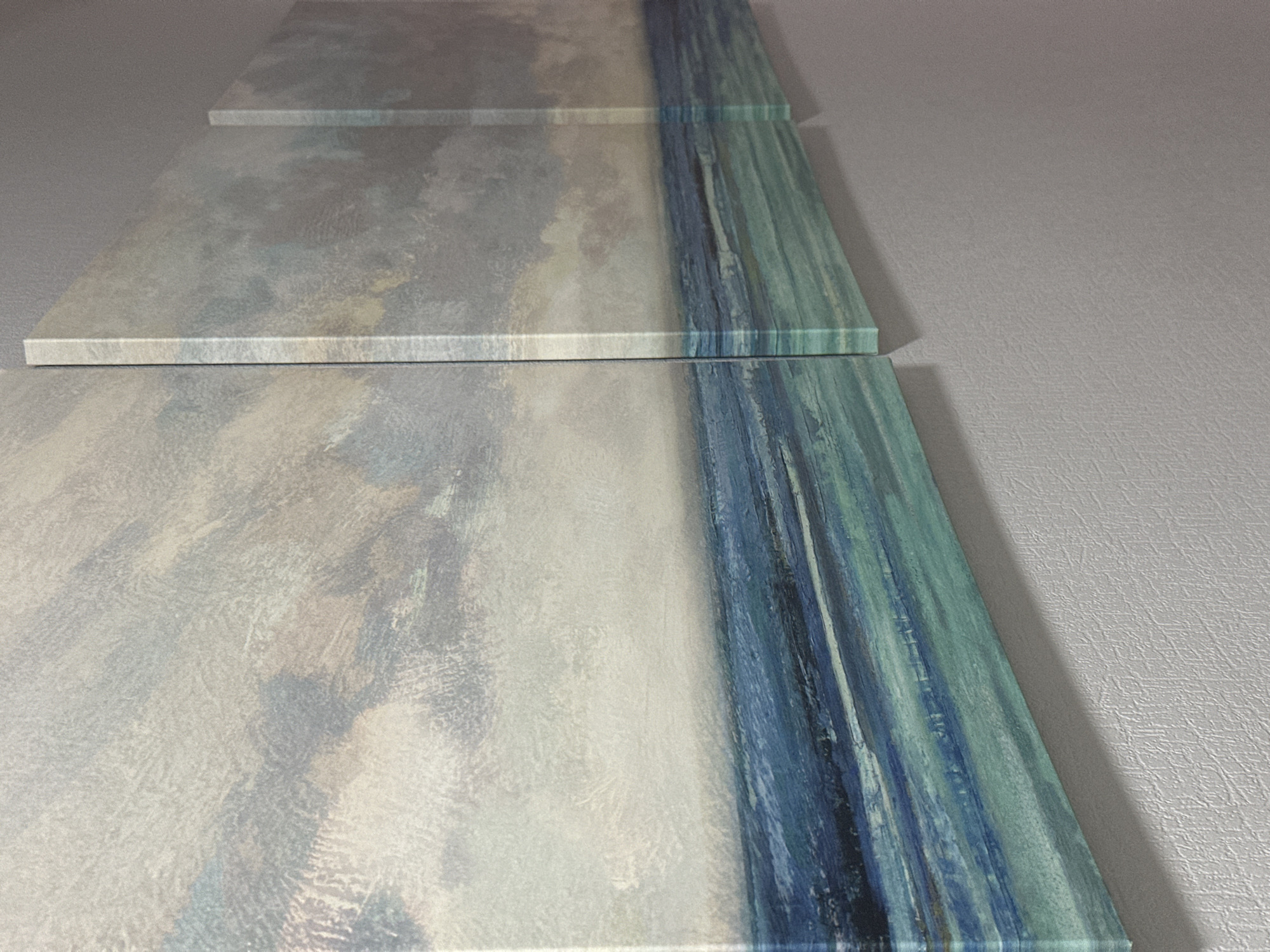 The image is a close-up, side-view photograph of a triptych canvas artwork depicting a seascape. The artwork is mounted on a wall and consists of three panels of varying widths, arranged horizontally. The leftmost panel, the widest, shows a textured sky with abstract brushstrokes in shades of white, grey, and muted blue. Below the sky is a horizon line separating it from the sea. The sea is depicted with horizontal bands of various blues and greens, also with a textured, impressionistic style. The middle and rightmost panels are narrower and continue the seascape motif, with the sky and sea textures matching those of the left panel. The artwork is hung on a light grey, textured wall. The lighting suggests an indoor environment, casting a subtle shadow on the wall from the artwork. There are no people, animals, or specific landmarks visible in the image. The location in the city of Nevinnomyssk, Russia is not discernible from the visual information provided in the image.