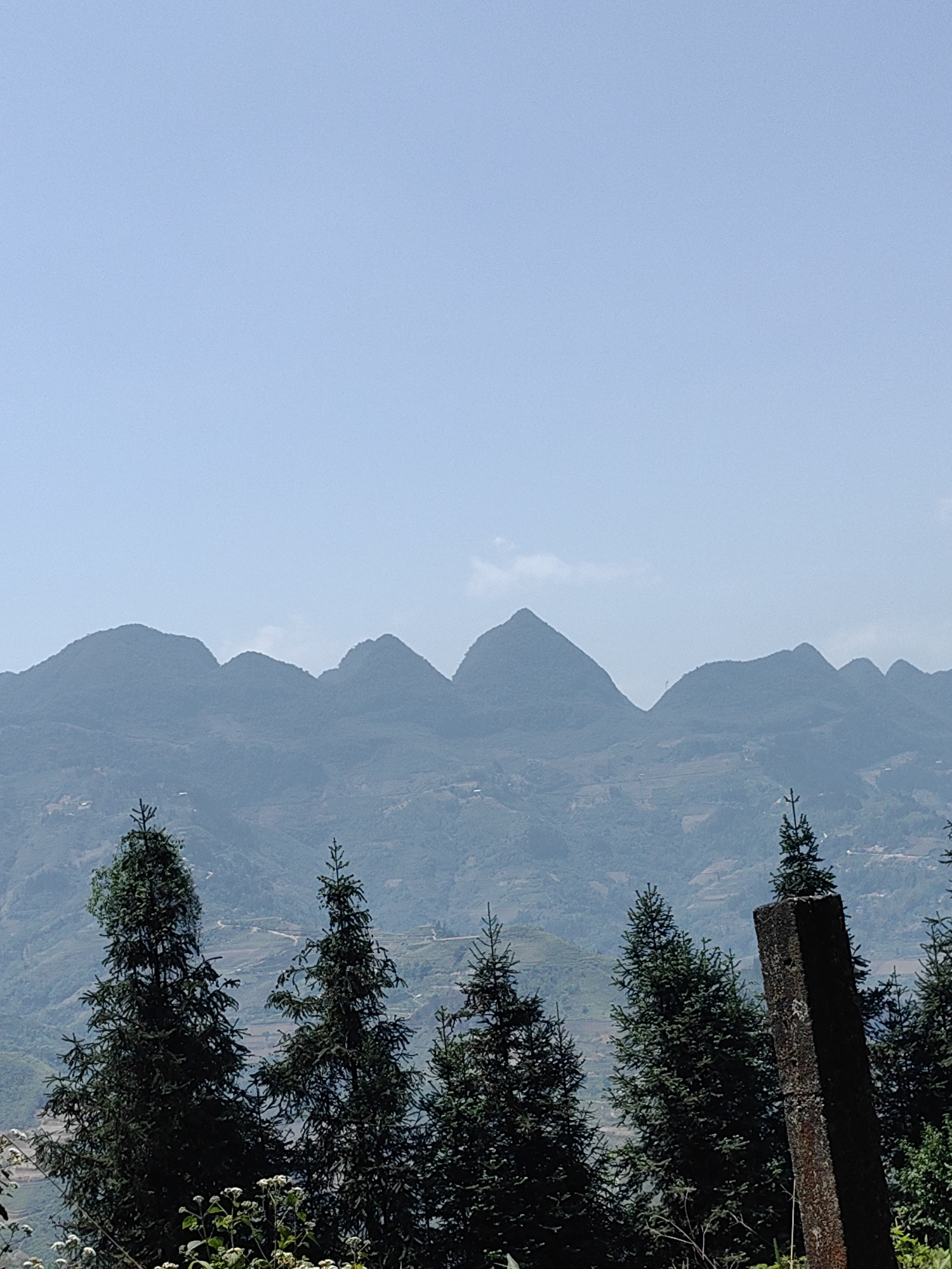 The image shows a mountainous landscape in Bạch Đích Commune, Vietnam. In the foreground, several evergreen trees are present. A weathered, rectangular stone pillar stands among the trees. The middle ground features a series of mountains with varying peaks, their slopes showing signs of terraced cultivation. The background is filled with a clear, light blue sky with some traces of clouds.