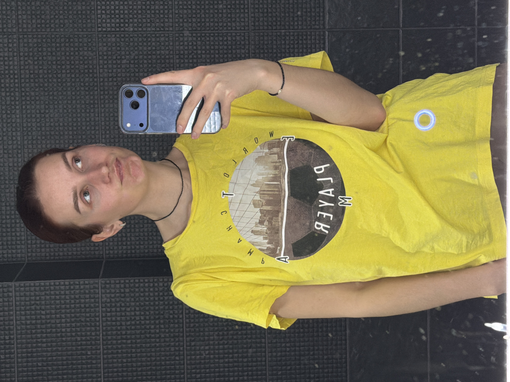 A woman is taking a selfie in what appears to be an indoor location with a gray tiled wall. She is holding a blue smartphone with a triple-lens camera. The woman is wearing a yellow t-shirt with a circular graphic containing a skyline and the reversed word "PLAYER." She is also wearing a black choker and a beaded bracelet. The location is presumably in the city of Chaikovsky, Russia, based on the prompt.