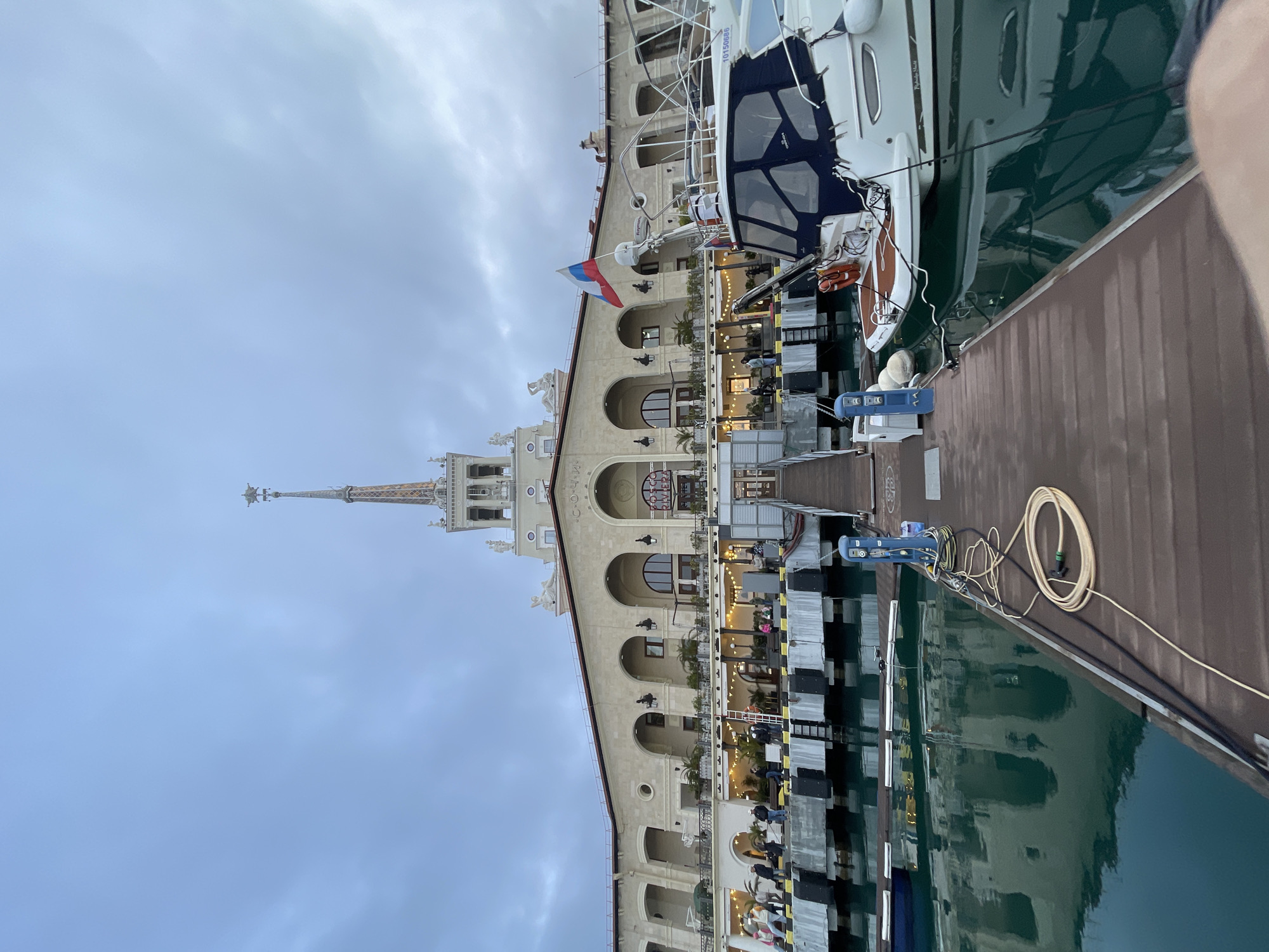 The image displays the Sochi Sea Port (Morskoy Vokzal) in Sochi, Russia, viewed from a wooden pier extending into the water. The multi-story port building, constructed in a Stalinist Empire architectural style, features a light-colored stone facade with numerous arched windows and balconies. A tall central spire, adorned with a star at its apex, dominates the skyline. The building's upper facade prominently displays the word "Cочи." Sculptures are visible on the building's upper levels and along the roofline.
On the lower levels of the building, outdoor dining areas are established on balconies, illuminated by string lights and decorated with potted plants. Multiple individuals are present on these balconies and along the dock structure, engaged in walking, standing, and dining.
In the immediate foreground, a dark brown wooden pier features mooring cleats, blue utility pedestals, a coiled yellow hose, and black electrical cables. To the right, a white motor yacht, equipped with a dark blue canvas canopy and an orange life buoy, is moored. A Russian national flag flies from the yacht's stern, and the hull number "10150686" is visible on its side. The calm, dark turquoise water reflects the building's facade. The sky above is overcast, a uniform light grey-blue, suggesting a low-light environment, likely late afternoon or early evening.