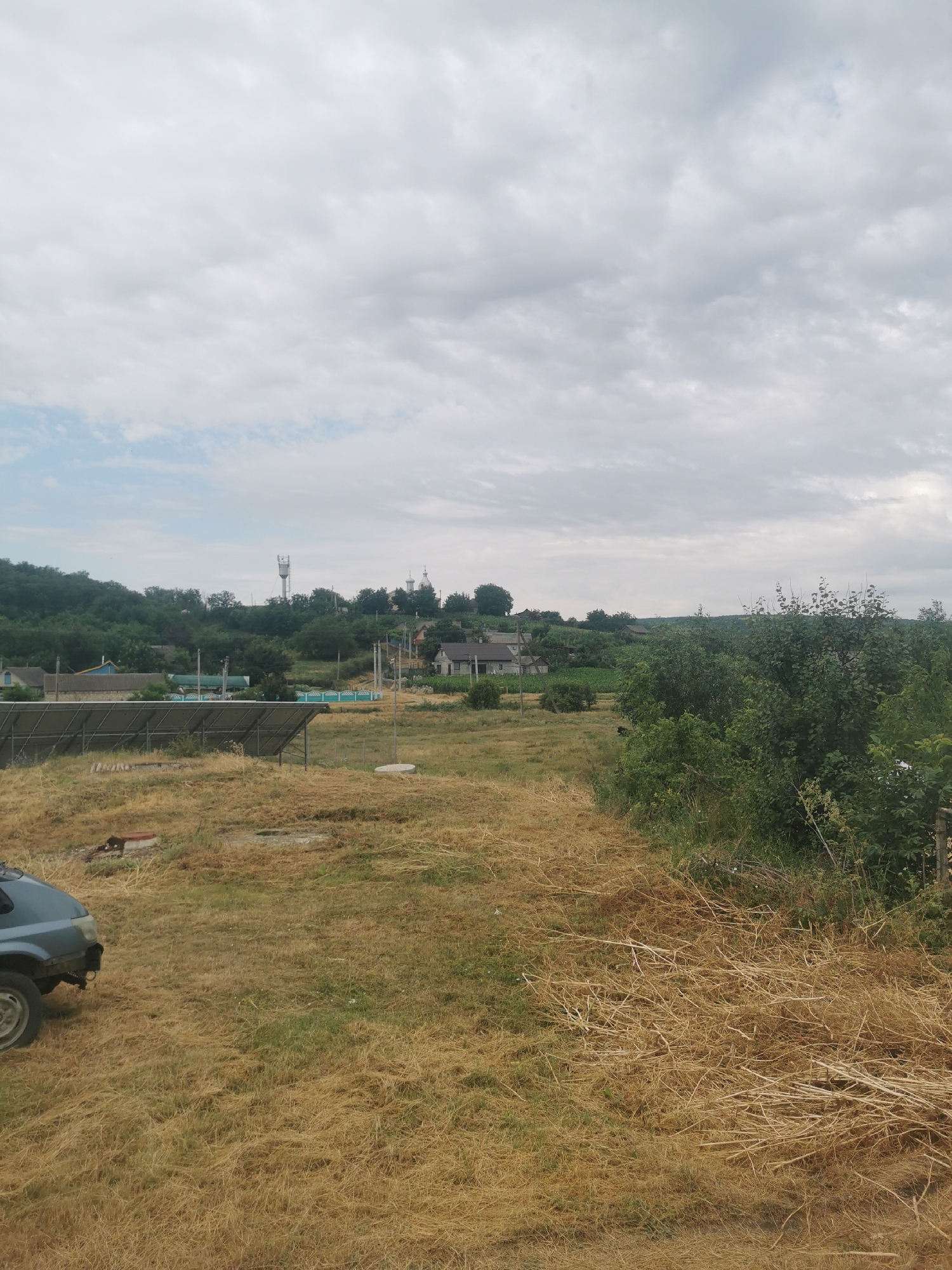 The image depicts an outdoor scene in the village of Carbună, Moldova. In the foreground, a portion of a blue car is visible on the left, parked on dry, light-brown grass. The terrain slopes upwards, revealing a field of dry grass and some scattered vegetation. A solar panel installation is situated in the middle ground, with several panels laid out on metal frames. Beyond the solar panels, a turquoise fence encloses a yard with some buildings. In the background, a cluster of houses and trees are nestled on a gentle hillside. A tall communication tower stands prominently on the horizon, and two domed structures, possibly churches, are visible further in the distance. The sky is overcast with scattered clouds. The overall impression is of a rural residential area on a cloudy day.