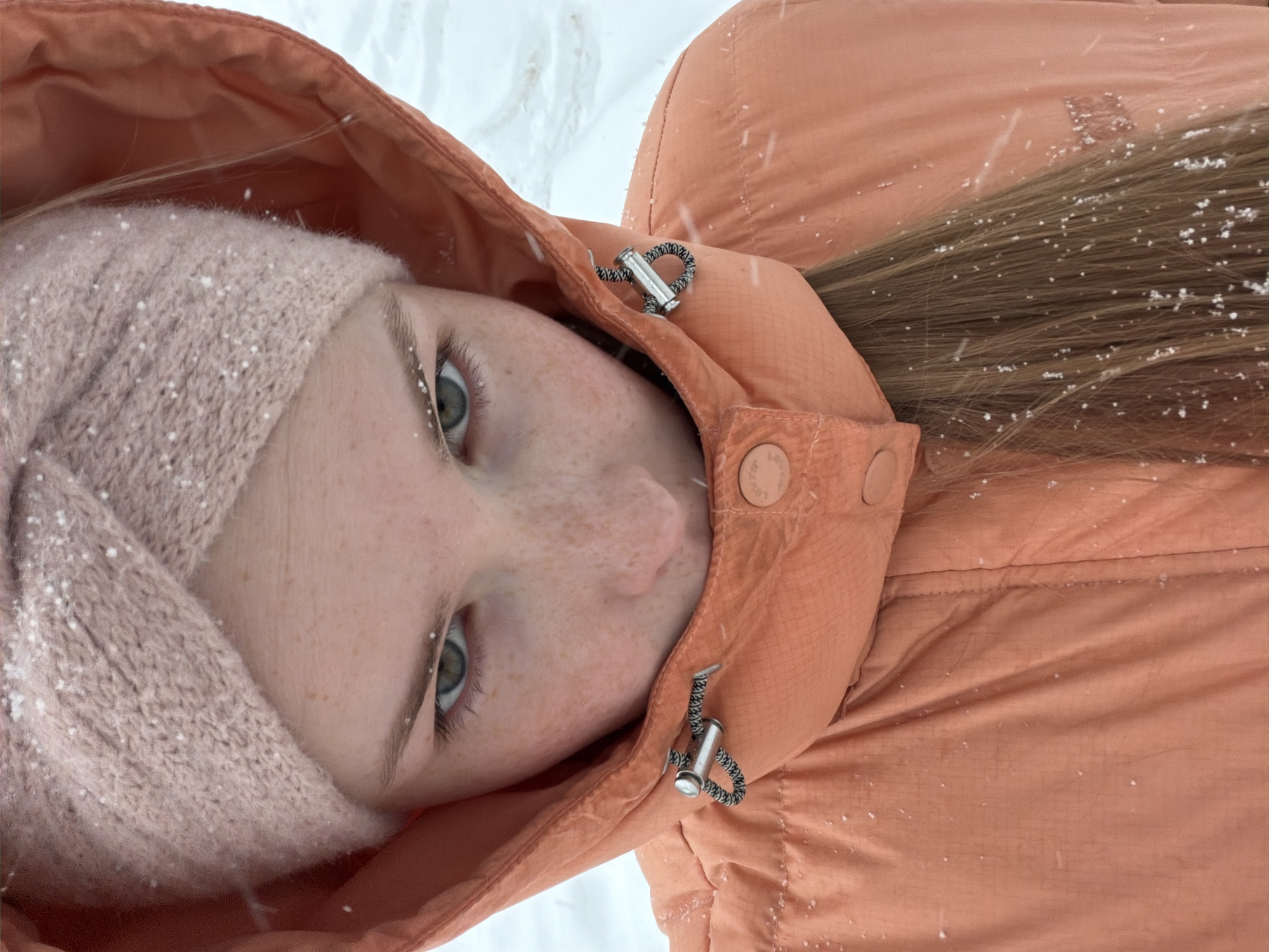 The image displays a close-up, top-down view of a young person lying in a snowy environment, likely in Berezniki, Russia. The subject, appearing to be female, looks directly at the camera with a serene and neutral expression. Her light green or hazel eyes are striking, complemented by numerous freckles across her nose and cheeks.
She is warmly dressed in a light peach or orange winter jacket, with the hood up. The jacket features visible drawstrings with silver-toned stoppers and circular buttons. Beneath the hood, she wears a light pink, knitted beanie or headband. Long, brown hair cascades from under the hood, resting on the jacket's fabric.
The background is an expanse of white, indicating snow-covered ground. Numerous small, white snowflakes are scattered across her jacket, hair, and knitted headwear, as well as on her eyelashes and eyebrows, suggesting that it is actively snowing. The lighting is bright and diffuse, consistent with an overcast daytime during winter. The photograph captures a quiet moment of enjoying the snowy weather. No visible text is present in the image.