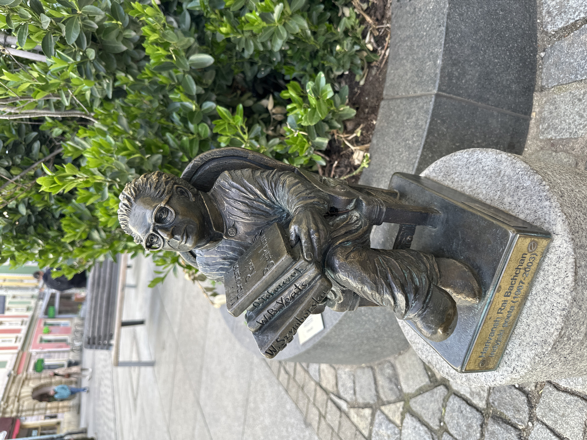 The image depicts a bronze sculpture of Harivansh Rai Bachchan, an Indian poet, located in Wrocław, Poland. The statue portrays Bachchan seated in a chair, holding a stack of books by other poets including C. Miłosz, W.B. Yeats, and W. Szymborska. The sculpture rests on a gray stone base, with a plaque inscribed with Bachchan's name and years of life (1907-2003). Behind the sculpture, there are green bushes. In the background, a blurred street scene with buildings and a bench is visible. The ground is covered with gray stone pavers.