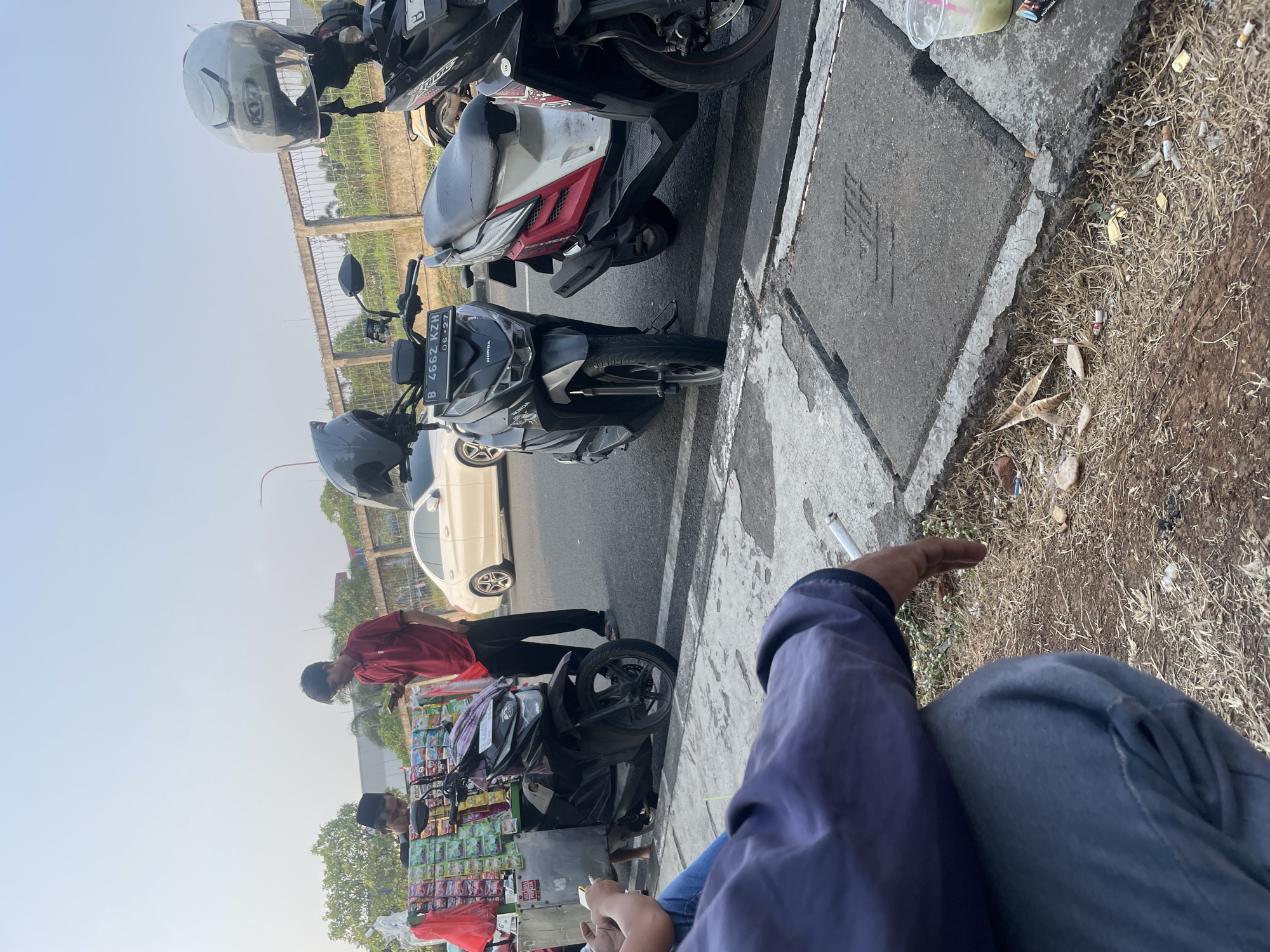 In Danauindah, Indonesia, a street scene unfolds by a road lined with a concrete curb and a grassy embankment. Two men are present. One man, wearing a red polo shirt and black pants, stands near a mobile food cart laden with various packaged snacks. He is holding a smartphone. Another man, wearing a black shirt and a skullcap, is behind the food cart, also appearing to be engaged with his phone or the cart's contents.
Three motorcycles are parked along the road. The closest motorcycle is a black scooter with a license plate displaying "B 4662 KZH" and "06-27". It has a gray helmet resting on its handlebars. To its right is a red, white, and black scooter, also with a helmet on its handlebars. Further to the right, partially visible, is another black motorcycle.
In the foreground, a person wearing a dark blue shirt and gray pants is seated on the ground, facing away from the viewer. They are holding a cigarette and a smartphone. The ground around them is covered with dry grass, litter, and cigarette butts. A clear plastic cup with a green beverage and a pink straw is visible on the curb to the right of the seated person. In the background, a white car drives on the road, and a chain-link fence with vegetation behind it runs along the side of the road. The sky is clear and bright, indicating daytime.