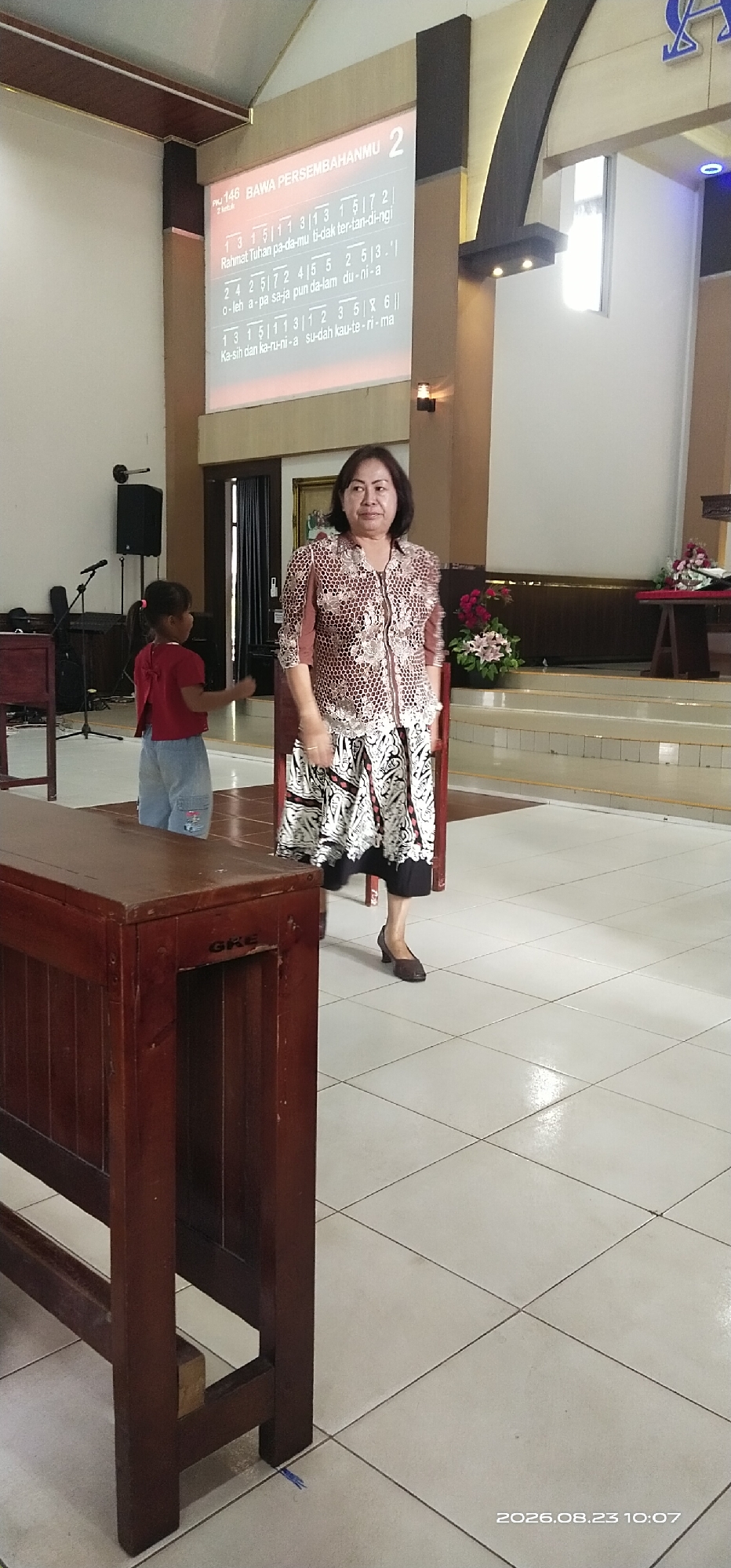 The image captures an indoor scene, likely a church, with a projector screen displaying lyrics and musical notation. A woman in a patterned blouse and a long, patterned skirt is standing and looking towards the right of the frame. To the left, a young girl in a red shirt and denim shorts is facing away from the camera, gesturing with her hands. In the foreground, a dark wooden pew or bench is visible. The environment features tiled flooring and a stage area with floral decorations in the background. The timestamp "2026.08.23 10:07" is present in the lower right corner. While the specific location in the city of Jaar, Indonesia, is not visually confirmed, the context of a church suggests a place of worship.