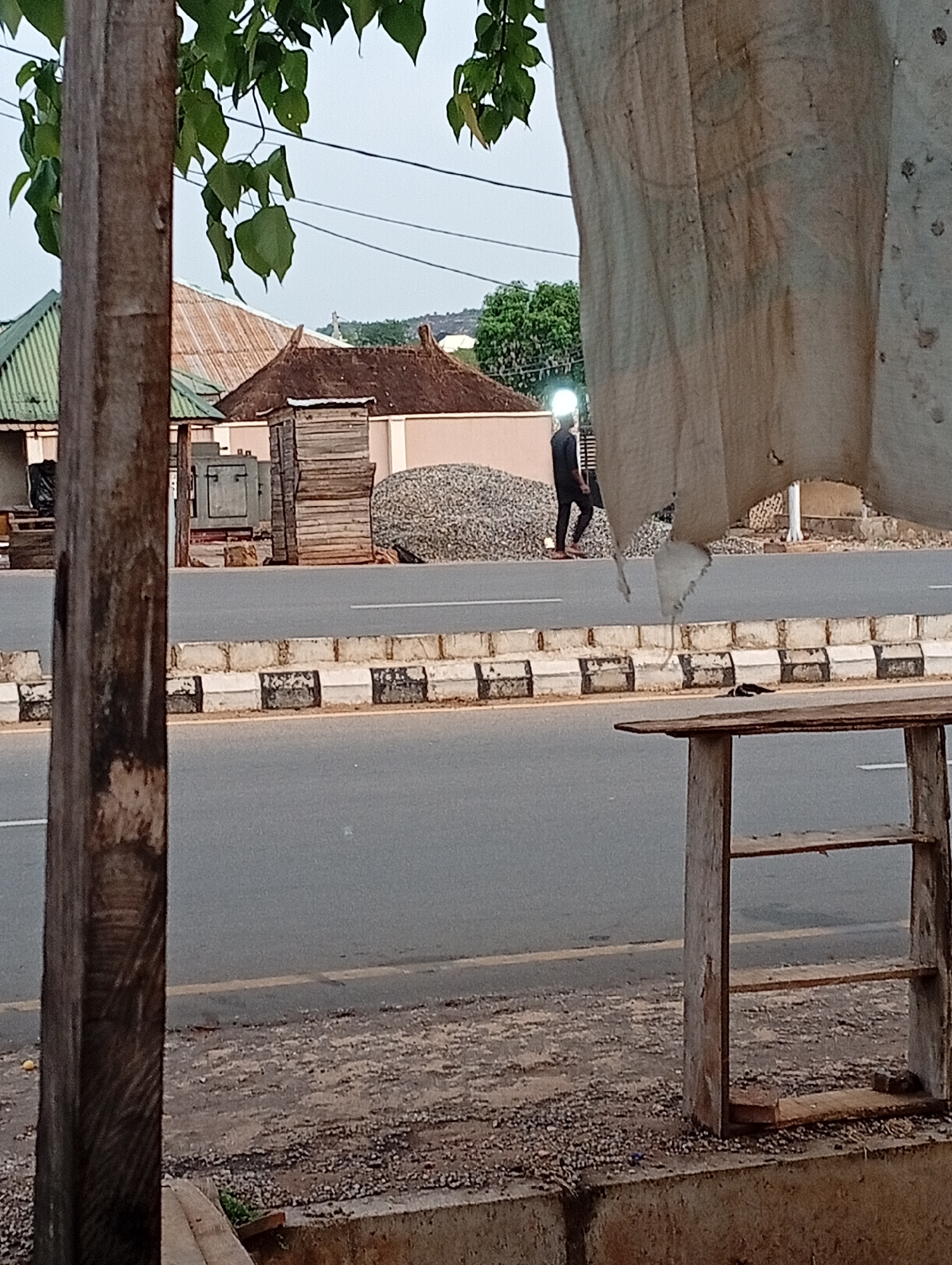 Adankolo, Nigeria. A man in a dark suit walks along the roadside next to a pile of gray stones. The road is divided by a curb painted with alternating black and white blocks. A wooden pole stands on the left and a tattered white cloth hangs on the right, partially obscuring the view. A wooden structure stands on the right side of the road. Buildings with varying roofs are visible in the background. The sky is overcast, and leaves hang from branches in the upper portion of the frame.