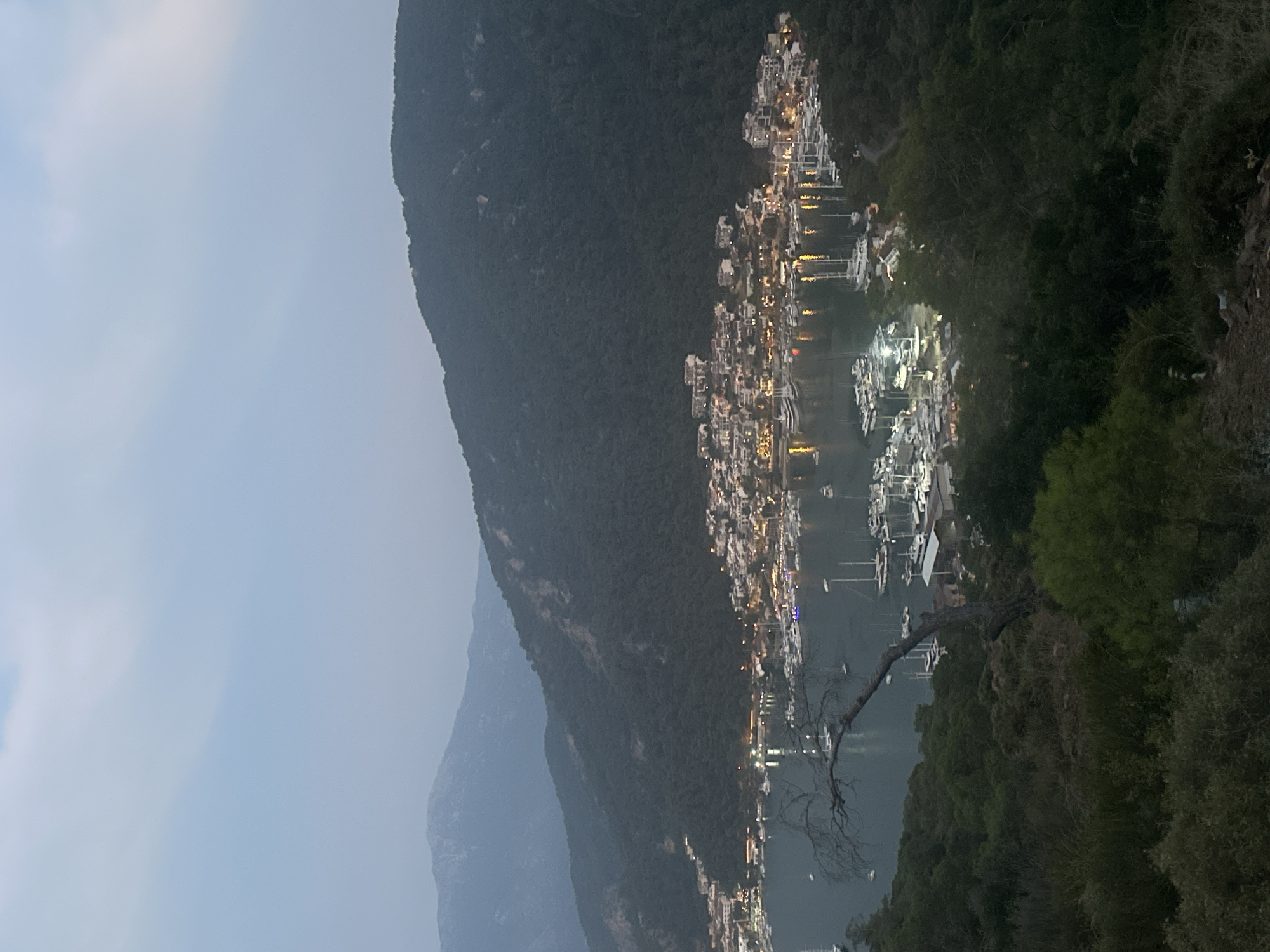 The image depicts a twilight view of a coastal town nestled at the base of a lush, tree-covered mountain range, likely in the vicinity of Fethiye, Turkey. A harbor filled with numerous sailboats and yachts occupies the foreground, their masts creating a sharp silhouette against the water. The town itself is illuminated with warm lights, suggesting evening or nightfall. Buildings are densely packed along the waterfront and extend up the gentle slope of the hill behind the harbor. The sky above is a soft, hazy blue with hints of clouds, and distant mountains fade into the atmospheric perspective. The overall scene conveys a sense of tranquility and a picturesque Mediterranean coastal setting. There are no people visible in the image.