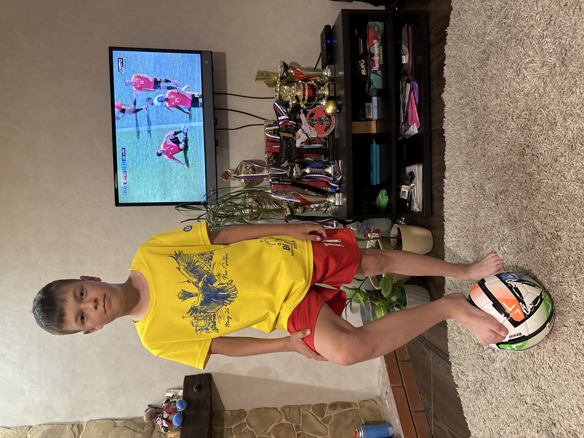 A young boy, likely around 10-12 years old, stands in a living room in Chaikovsky, Russia. He is wearing a yellow t-shirt with a graphic of a bird and buildings, and red shorts. He is holding a soccer ball with his feet, which is placed on a beige carpet. To his left, a television displays a soccer match with players in red and white uniforms. Behind the television, a black shelf unit is filled with numerous trophies and some boxed games. The room appears to be lit by artificial light, suggesting it is either evening or daytime with the curtains drawn. The overall mood is casual and domestic, with the boy engaged in a playful activity. Visible text includes "RESIST" on the soccer ball and various labels on the games and TV screen, though they are too small to read clearly.