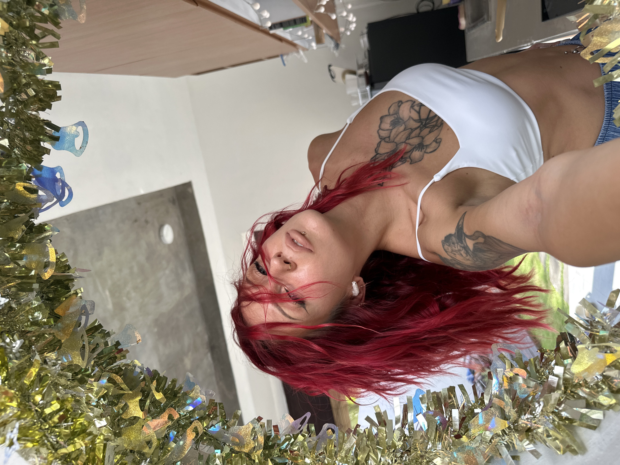 The image shows a woman with red hair, wearing a white top, with tattoos on her chest and arm. She is framed by gold and blue holiday tinsel. The background includes a grey wall, a white wall with a door, and a television. The image appears to have been taken in North Kuta, Indonesia.