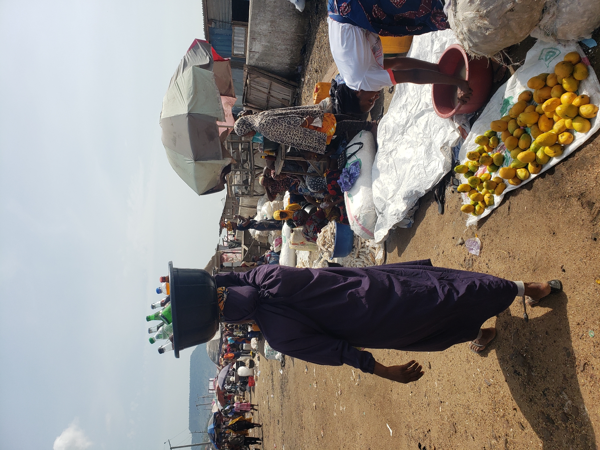 A busy market in Lokoja, Nigeria. The foreground shows a person balancing a black bucket full of glass bottles on their head. In the middle ground, numerous vendors sit under umbrellas, selling various goods. To the right, a person is washing something in a red bucket next to a pile of yellow fruits. The background contains a mountain range under a bright sky.