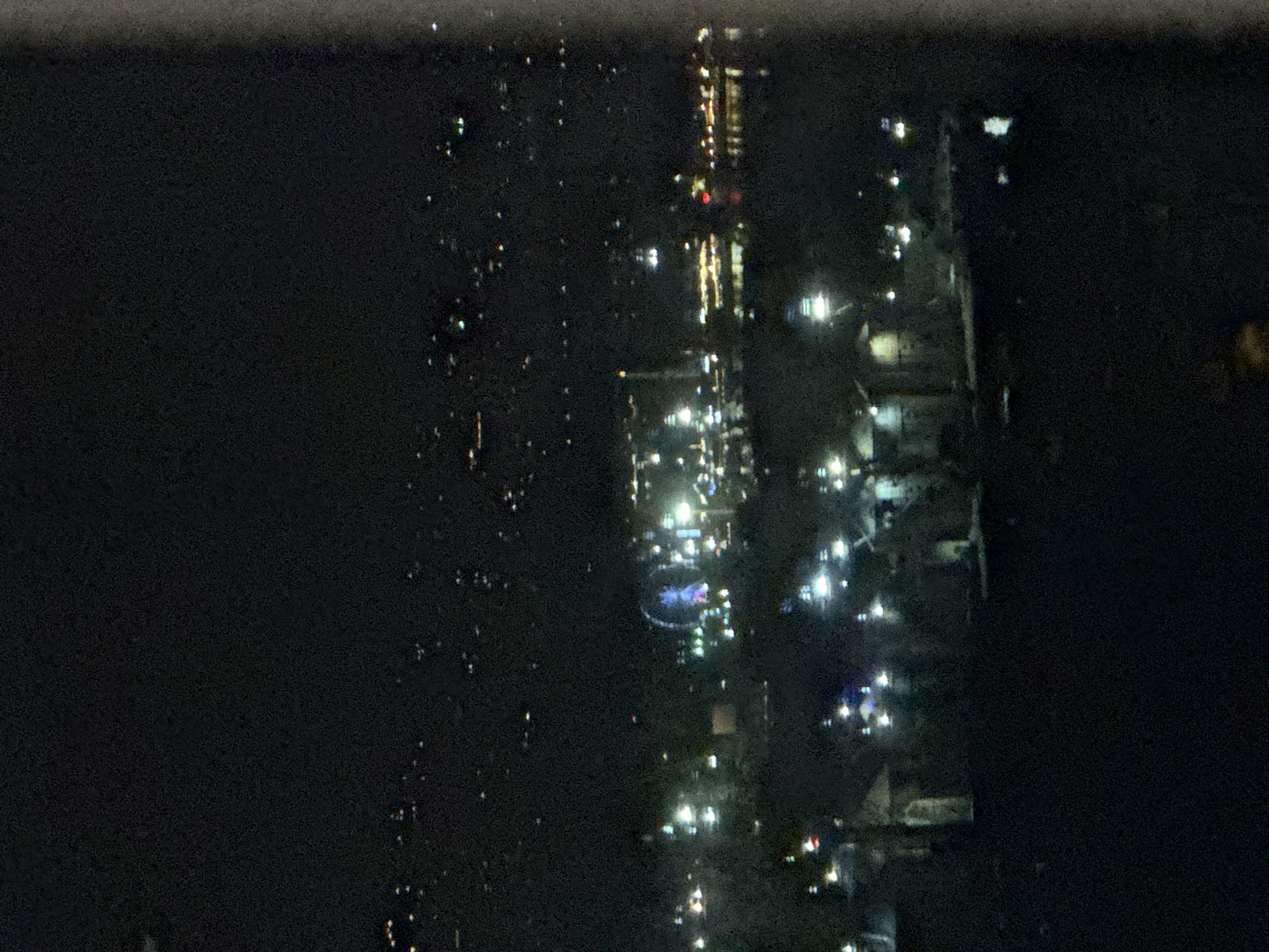 An aerial night time photograph shows a blurred cityscape with a Ferris wheel illuminated in blue and pink lights. The cityscape is dotted with numerous small lights, suggesting buildings and streetlights spread across the area. In the foreground, there is a darker, blurry area, possibly representing buildings or structures closer to the viewer. The overall image is dark with a significant amount of digital noise. The scene appears to be a well-lit entertainment area or a section of a city at night.