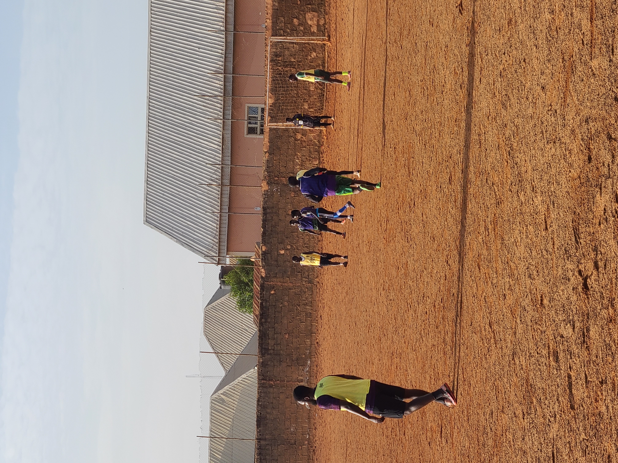 The image depicts a group of individuals, presumably soccer players, training on a red dirt field in Jos, Nigeria. The players are wearing assorted training bibs over shirts, some in yellow and purple, others in blue. They appear to be performing warm-up exercises. The field is bordered by a brick wall and buildings with corrugated metal roofs. A soccer goal can be seen along the brick wall. The sky is light blue, suggesting daytime.