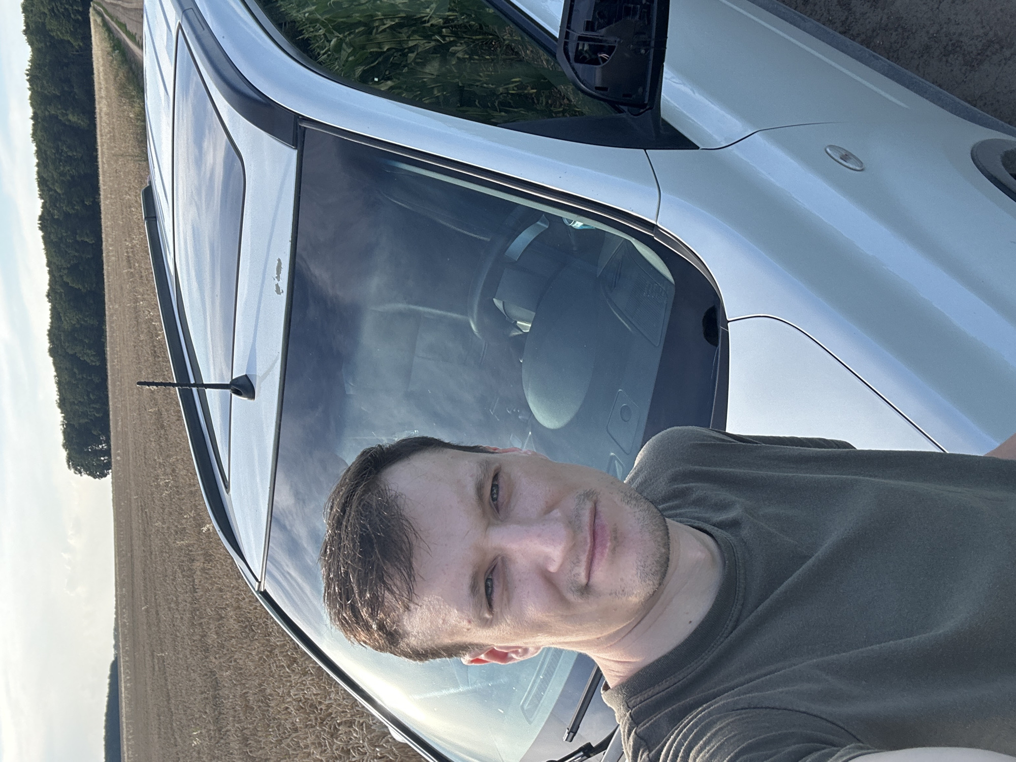 A medium shot selfie shows a man standing in front of a white vehicle parked on a dirt road. He is wearing a dark green t-shirt and has short brown hair and a mustache. The vehicle has a sunroof and an antenna on the roof. The background consists of a field of dry grass, a line of trees, and a cloudy sky. The setting appears to be a rural area, possibly near Makiv, Ukraine.