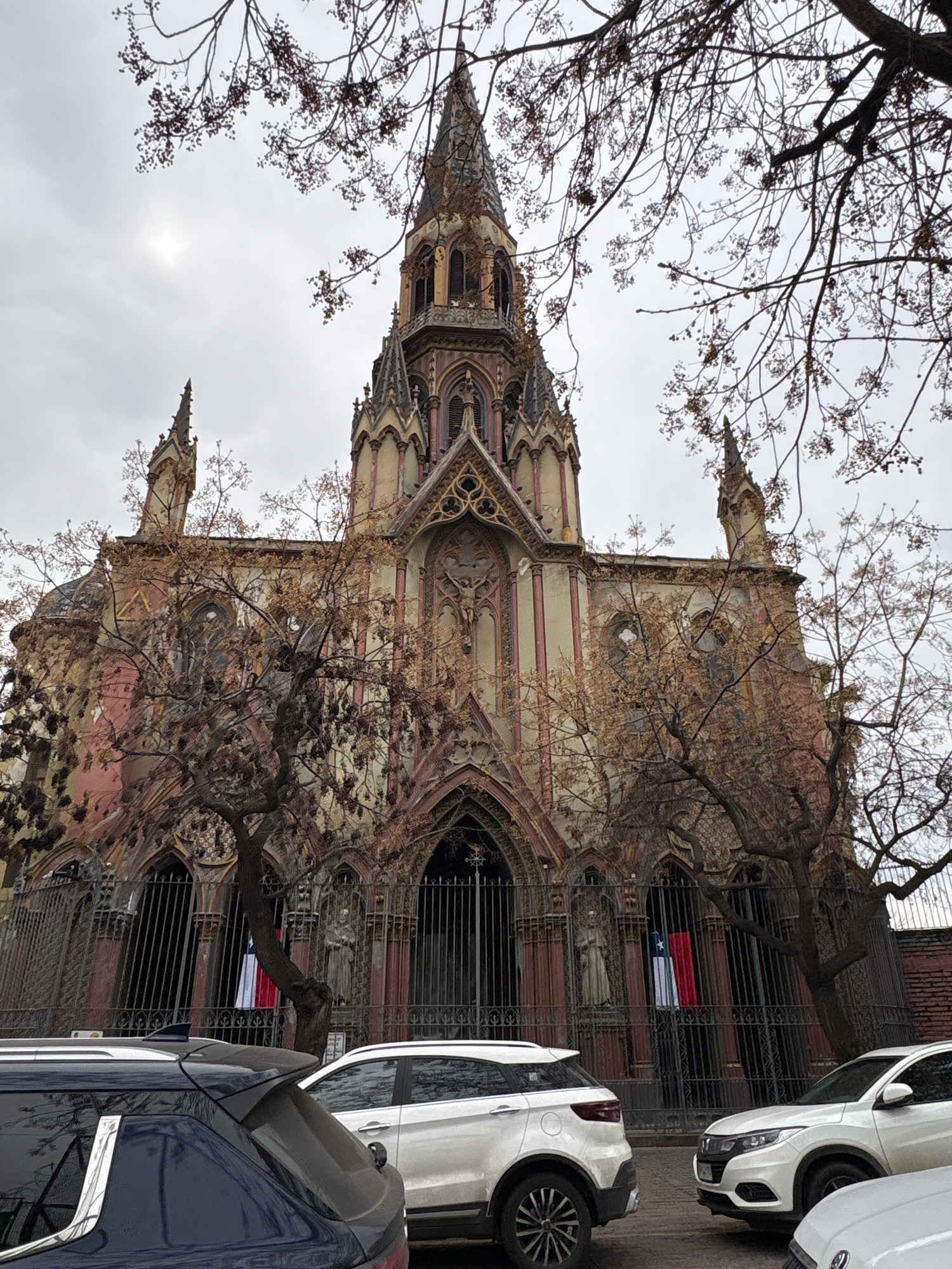 In Santiago, Chile, a Gothic-style church, possibly the Basílica de la Merced, stands under a cloudy sky, its weathered facade displaying a mix of pink and beige tones with ornate architectural details. Bare tree branches, devoid of leaves, partially obscure the upper portion of the building and its prominent spires. The church is fronted by a dark metal fence, behind which two flags, one red and white with a blue star on a white rectangle, and another entirely red and white, are displayed, likely the Chilean flag. In the foreground, the rear of a dark blue SUV and the front of a white SUV are visible, indicating the presence of vehicles on a street in front of the church. The scene suggests a city environment, likely in the downtown area of Santiago, given the architectural style and urban elements.
