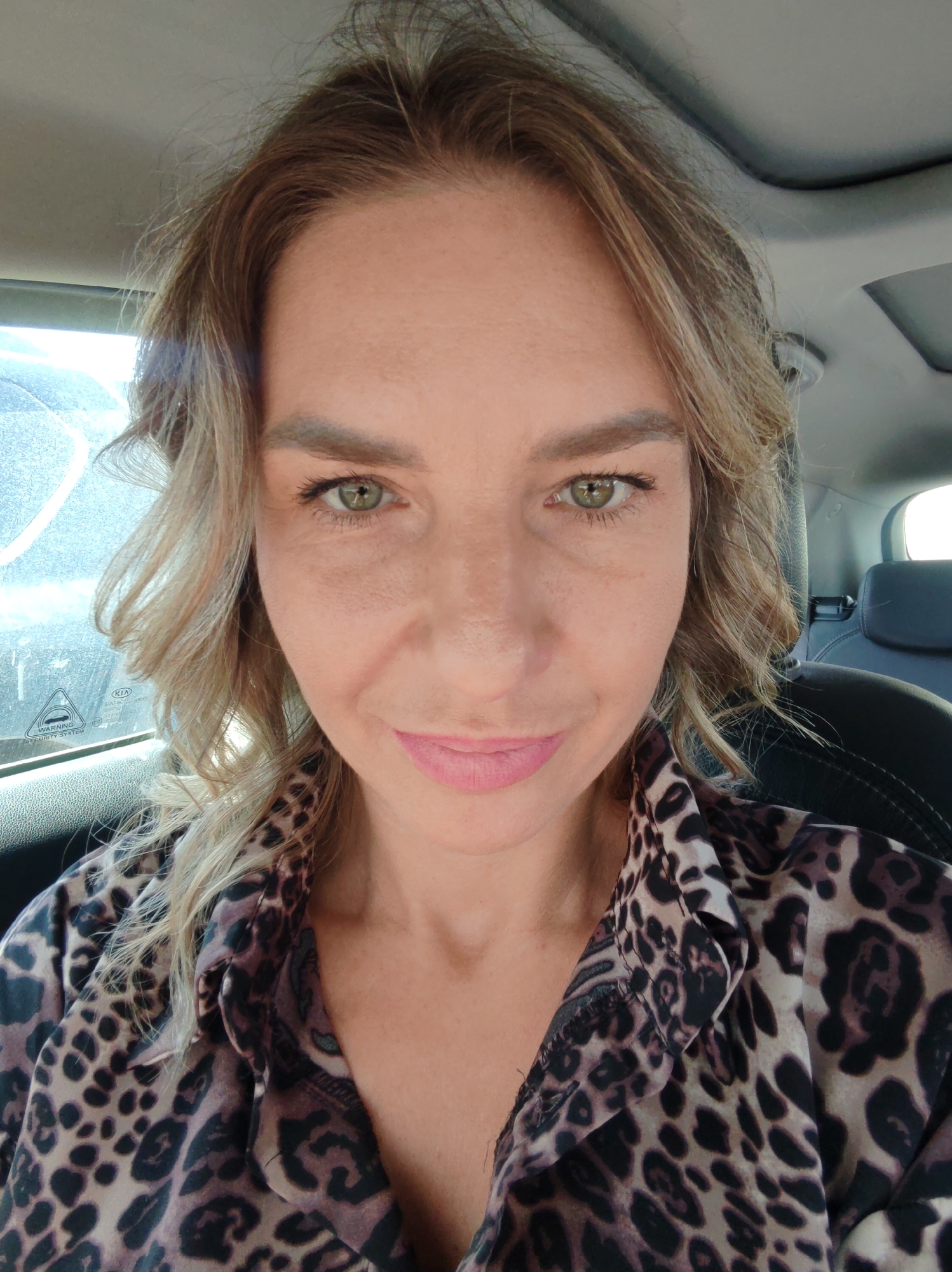 A woman is depicted in a close-up, front-facing portrait taken inside a car, likely in Petropavlovsk-Kamchatsky, Russia. She has light brown hair with blonde highlights, styled in soft waves, and piercing green eyes. Her eyebrows are well-defined, and she wears light makeup, including pink lipstick. She is dressed in a distinctive brown and black leopard print blouse.
The scene is set within the interior of a vehicle, with parts of a car seat, headrest, and the ceiling visible behind her. Bright daylight illuminates the scene, suggesting it is daytime. Light reflects on the car window behind her, which appears somewhat streaked or dusty. On this window, a sticker is partially visible, displaying the text "KIA WARNING SECURITY SYSTEM" along with some other indistinct characters.
The woman looks directly at the camera with a subtle, pleasant smile, conveying a calm and confident emotion. The activity appears to be a selfie or a casual portrait taken while she is a passenger or stationary within the vehicle.