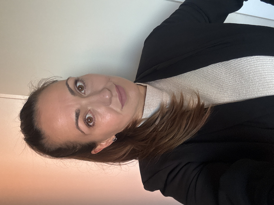 The scene depicts a medium close-up portrait of an adult female, situated in Varna, Bulgaria. The subject has brown hair, pulled back into a ponytail with some strands falling over her right shoulder. Her eyes are brown, and she has a fair complexion with light makeup, including eyeliner and lip color. She is attired in a black open blazer or cardigan worn over a light-colored, ribbed turtleneck top with a subtle shimmer. A small, dark-colored stud earring is present in her left ear. The subject is looking directly at the camera with a slight, closed-mouth smile. Her head is slightly tilted to her left. The background is a plain, light-colored wall, divided by a vertical seam; the section to the subject's left has a subtle peachy tint, while the section to her right is off-white or pale grey. The lighting is uniform, indicating an indoor environment.