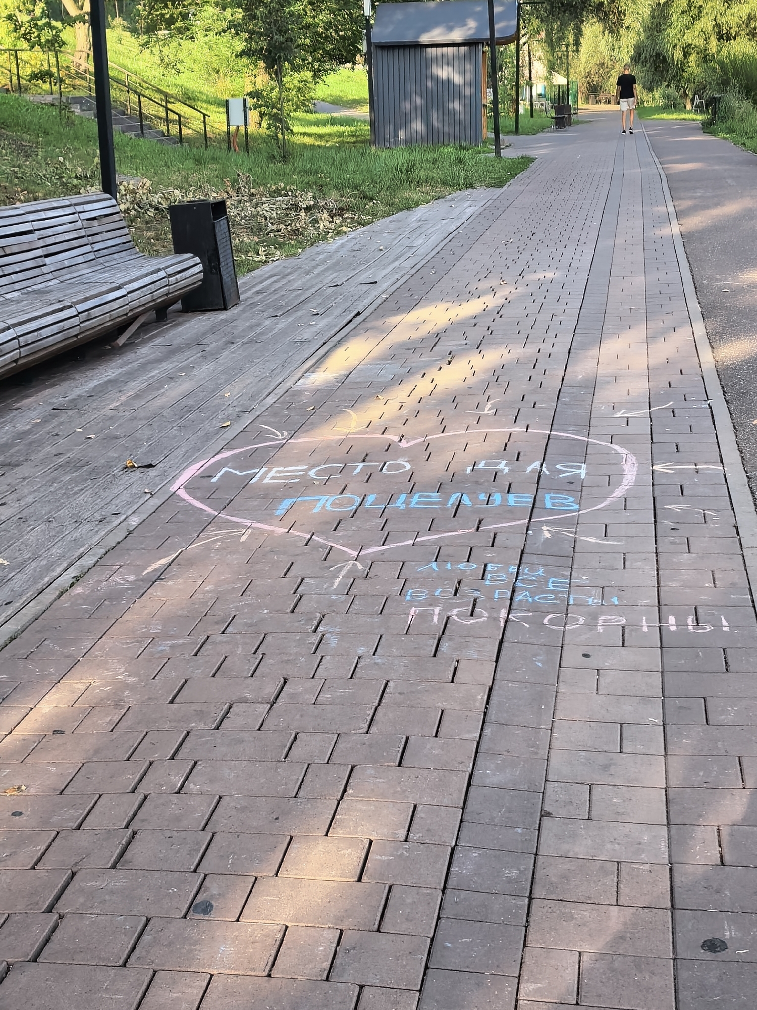 The image shows a paved pathway in a park, likely in Sterlitamak, Russia, based on the chalk inscriptions. The pathway is made of rectangular brick pavers. On the right side of the path, a person wearing a black t-shirt and light-colored shorts is walking away from the camera. To the left of the pathway, there is a wooden bench and a trash bin. Green grass and trees are visible in the background.
Chalk drawings are present on the pathway. A large pink heart encloses blue text. The visible text within the heart appears to be "МЕСТО ДЛЯ ЦЕЛЯЕВ" which translates to "A place for targets" or "A place for goals." Below the heart, in smaller blue text, is "ЛЮБВИ ВСЕ БЕГ", which translates to "love of all running" or "love of everyone running". Further down, more blue chalk text reads "ВСЁ ВПРАСТИ ПОКОРНЫ!" which translates to "Everything is simple and submissive!". There are also chalk arrows pointing in different directions. The sun casts long shadows across the pathway, indicating it is late afternoon or early morning.