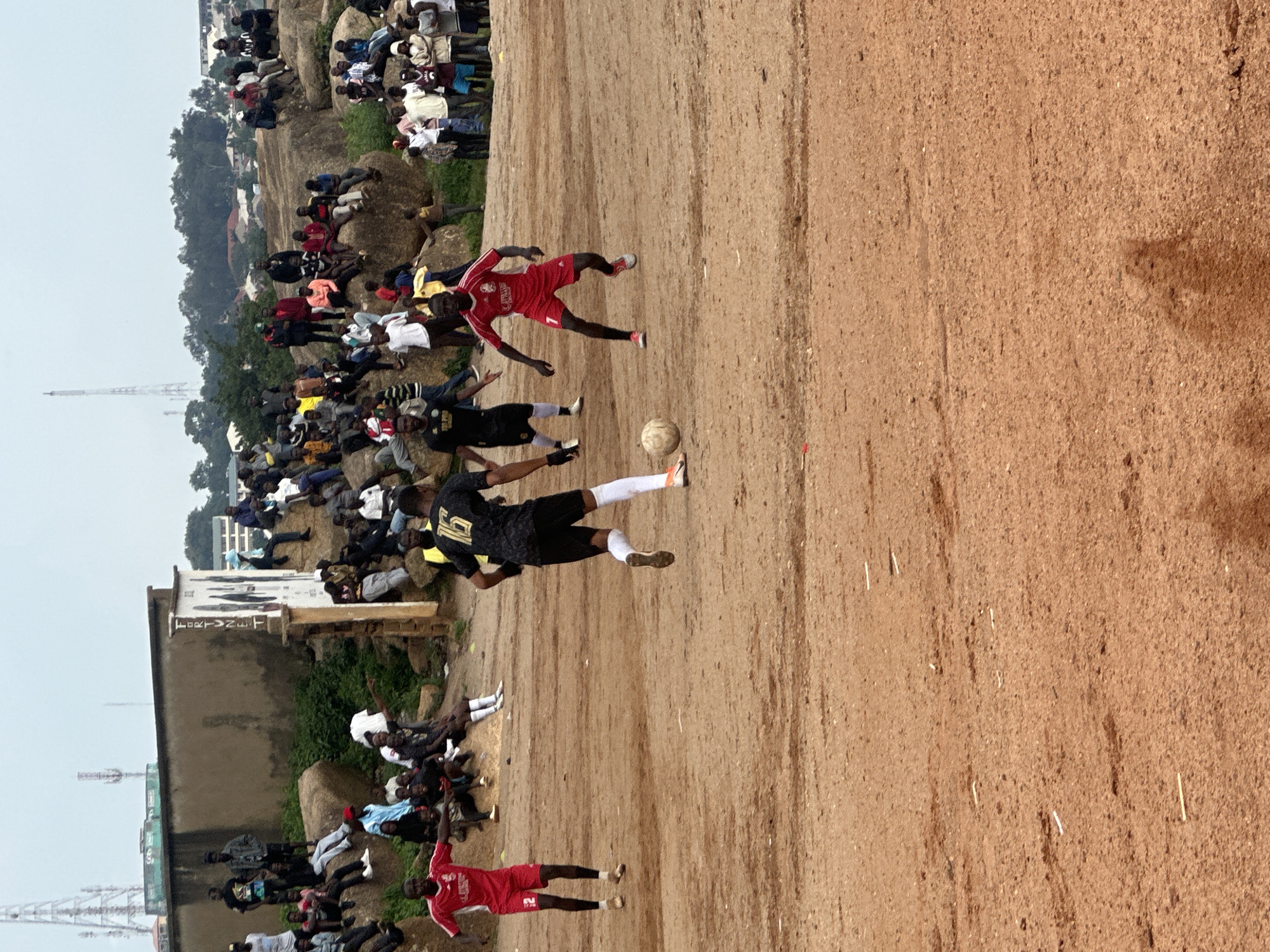 This is a vertical, medium-shot photograph of a soccer game taking place on a dirt field, with a large crowd of spectators gathered on rocky, elevated ground to the left of the field. The game appears to be taking place in Jos, Nigeria. Two players are visible in action on the field, one in a red jersey with the number 7 and white shorts, and another in a black jersey with the number 16 and black shorts. A soccer ball is visible near the player in the black jersey.
The spectators are densely packed, sitting and standing on the rocks and natural inclines, all looking towards the game. Many of the spectators are dressed casually in t-shirts and shorts. The background shows a mix of trees and buildings, with some radio towers visible in the distance, characteristic of an urban or semi-urban environment. The lighting suggests it is daytime, likely late afternoon, given the shadows cast on the dirt field. The overall atmosphere appears to be one of casual sporting enjoyment, with the spectators engaged in watching the match. There is no visible text in the immediate vicinity of the players or the ball.