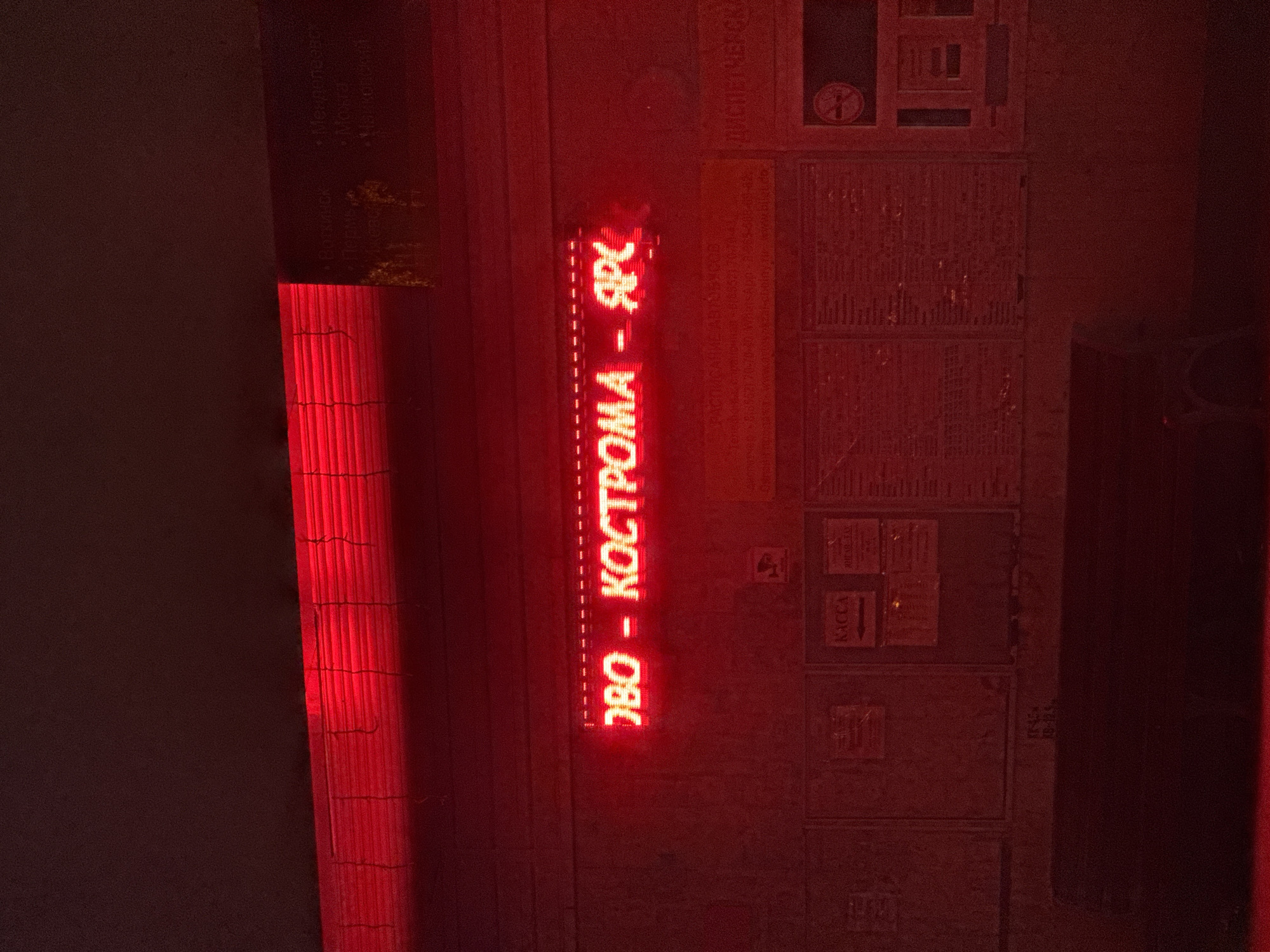 An exterior shot at night, bathed in red light, shows a digital sign displaying destination information. The sign reads "ЗБО - КОСТРОМА - ЯРС". Below the sign are several display boards with text and images, including one labeled "КАССА" (KASSA), which translates to "TICKETING OFFICE". The environment suggests a public transportation hub, possibly a bus or train station, given the destinations and the "KASSA" sign. The overall scene is dimly lit, with the red neon glow dominating the visual field.