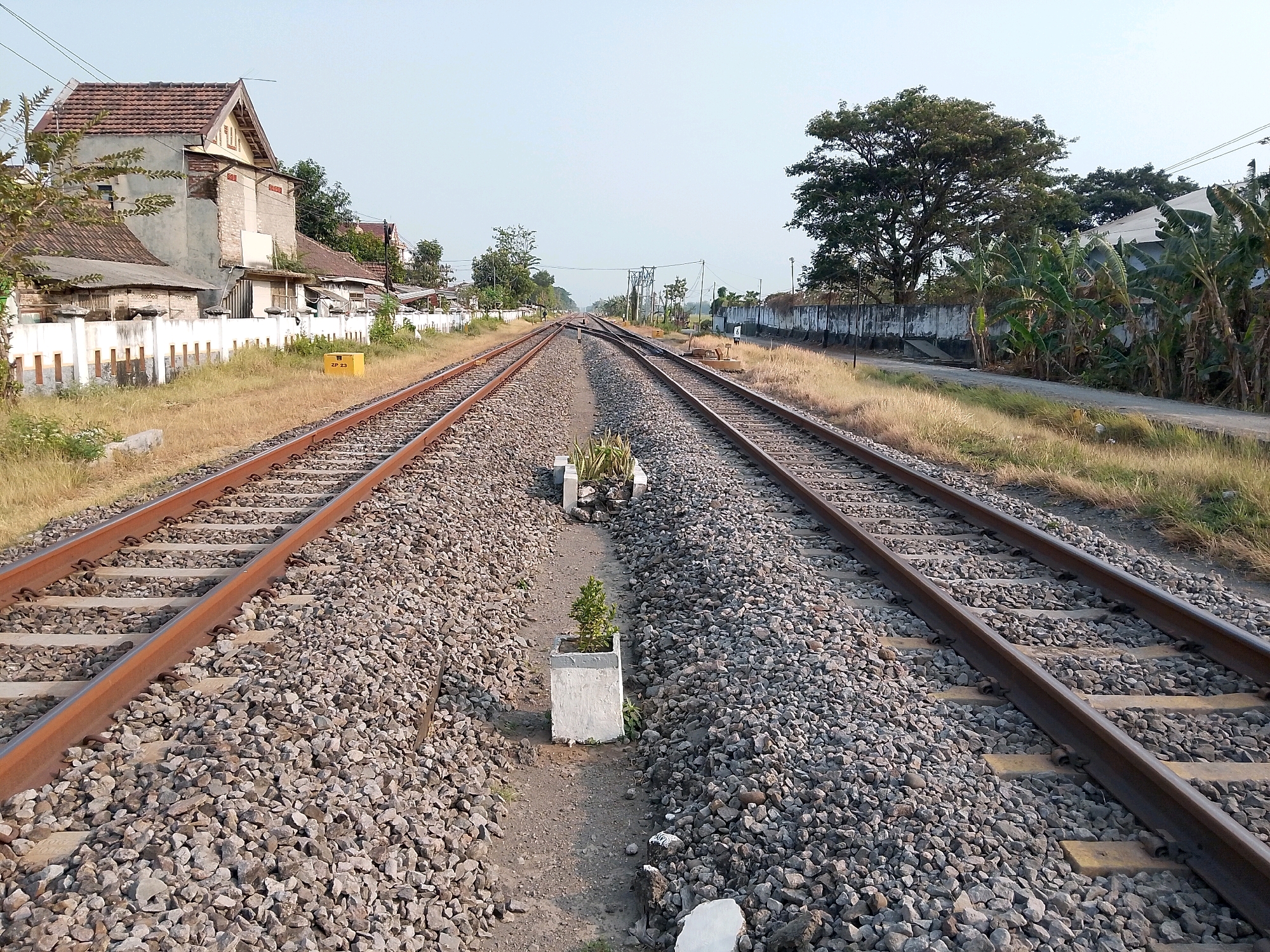 The image shows a double railroad track in Geneng, Indonesia, with a building and vegetation in the background. The tracks are made of steel rails laid on wooden sleepers, covered with gravel. The building on the left appears to be a house with a tiled roof and a white fence. The vegetation on the right includes banana trees and a large tree. The sky is clear and the lighting suggests it is daytime.