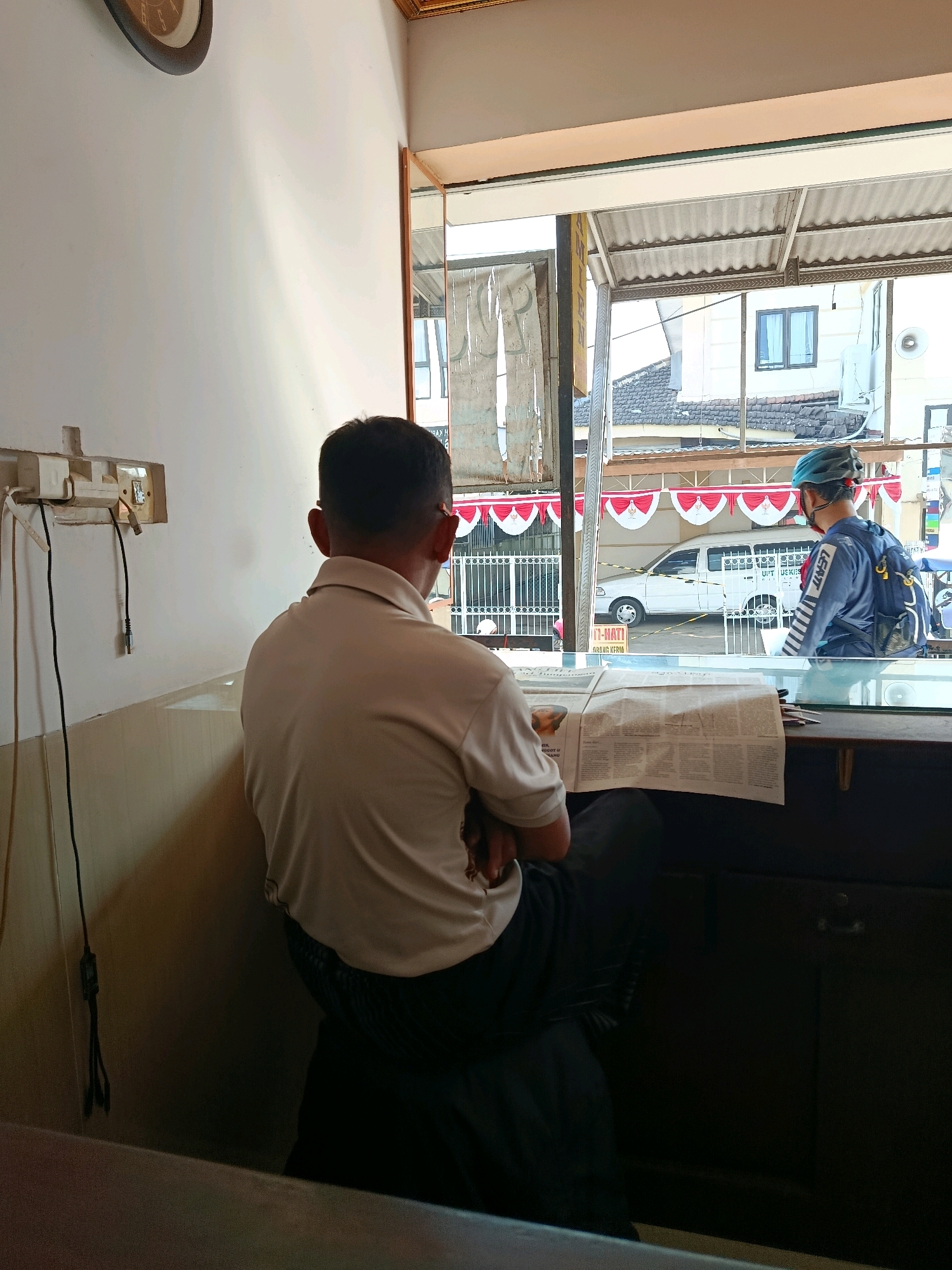 A man wearing a light-colored polo shirt and dark pants is seated facing away from the camera, reading a newspaper. He is sitting at a counter with a newspaper spread out before him. To his right, another man wearing a blue jacket and a blue helmet with a backpack is standing and looking out of a window. The background visible through the window shows a street scene with buildings, a white van, and decorative red and white banners. The interior setting appears to be a shop or cafe. The location is identified as Bululawang, Indonesia.