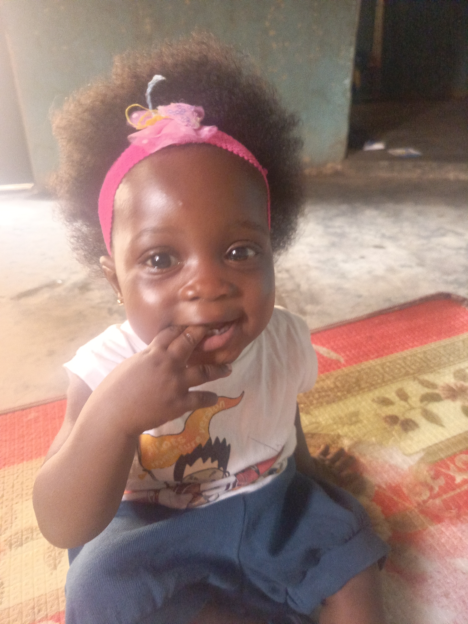 A close-up shot shows a young child sitting on a patterned mat in what appears to be a home in Akwanga, Nigeria. The child, wearing a pink headband in her afro hair, has a white t-shirt with a cartoon graphic and dark blue shorts. She has her fingers in her mouth and appears to be smiling. The background shows a wall with patchy paint. The mat is multicolored and appears to be made of woven fibers. The lighting is somewhat dim and appears to be natural light.