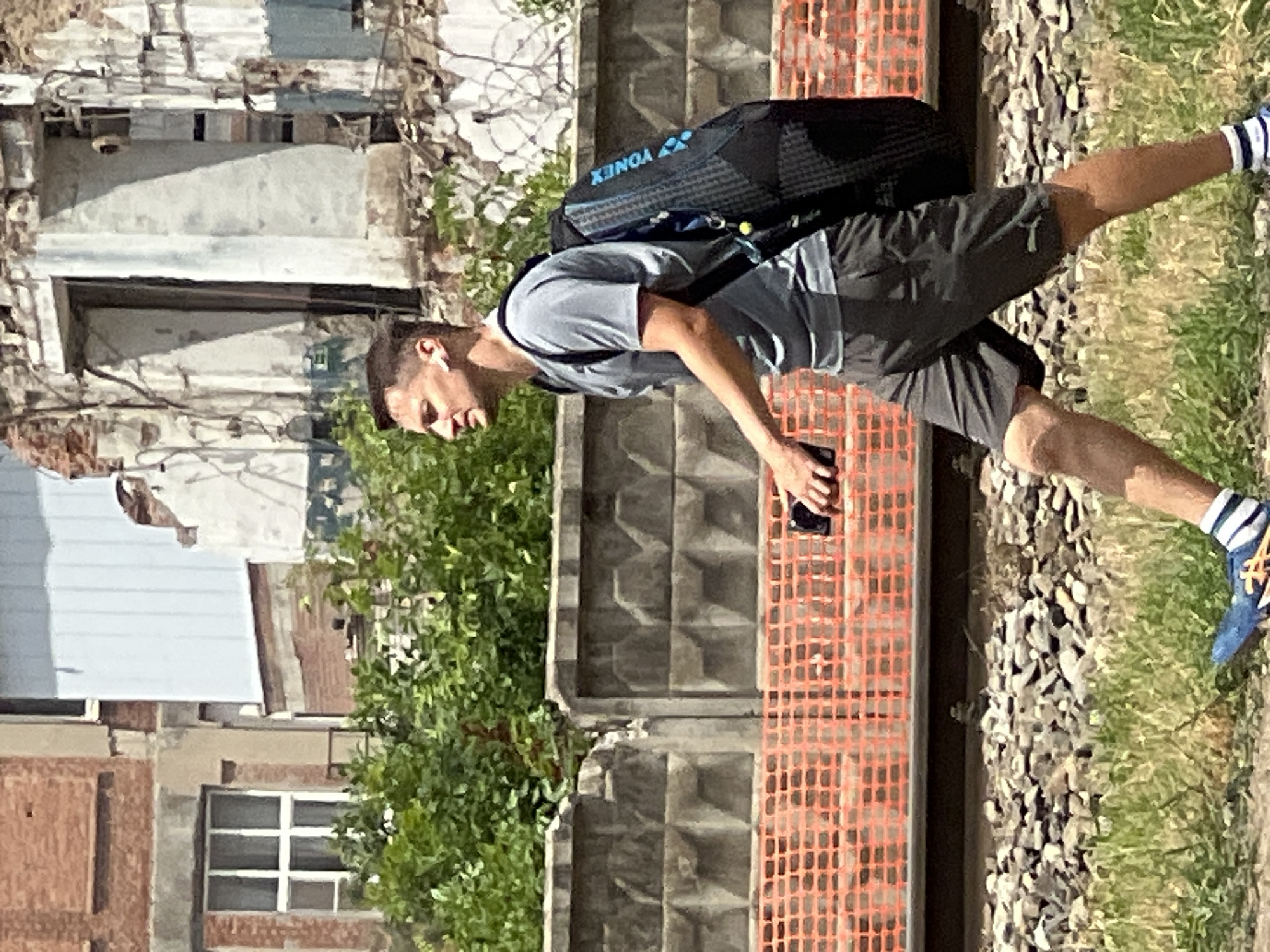 A man is walking along a railway line with a large backpack on. He is wearing a grey t-shirt, dark shorts, and blue athletic shoes with white socks. The man has short brown hair and is wearing white earbuds. He is holding a black smartphone in his right hand. The backpack is black with blue accents and has the Yonex logo on it. The background shows a damaged building with brick and concrete construction, overgrown vegetation, and a concrete fence. An orange safety net is visible in the foreground. The scene appears to be in an urban industrial area.