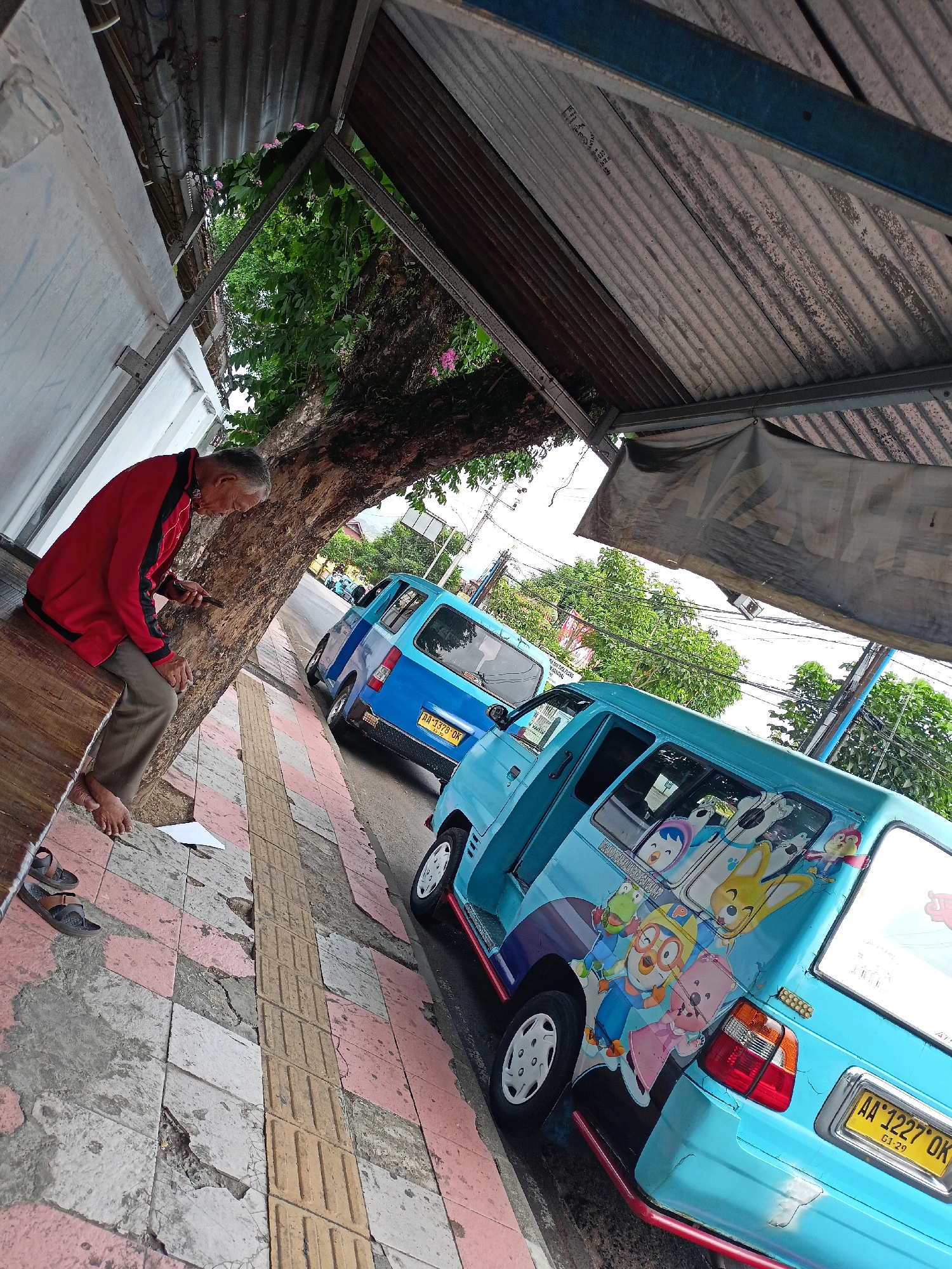 An elderly man wearing a red jacket and tan pants is sitting on a wooden bench next to a tree on a sidewalk in Magelang, Indonesia. He is looking down at his phone. Two light blue vans are parked on the side of the street. The van closest to the camera has cartoon characters on its side. The sidewalk is made of cracked tiles and a tactile paving strip. The roof of a structure, made of corrugated metal, is overhead.